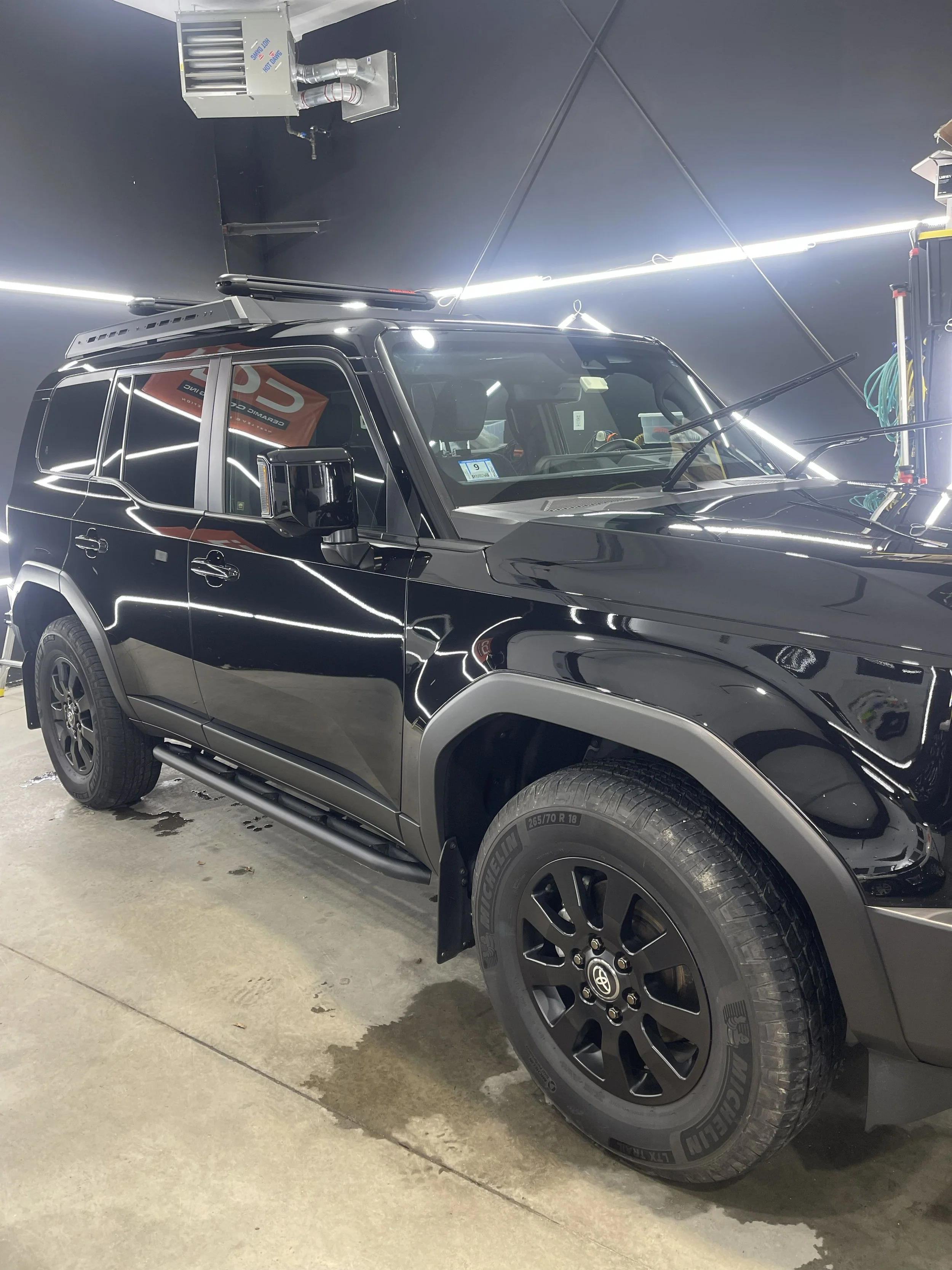 A black SUV parked indoors, with a roof rack, side mirrors, and tires visible. The vehicle is reflected in the shiny black paint, with a corner of the garage visible on the ground.