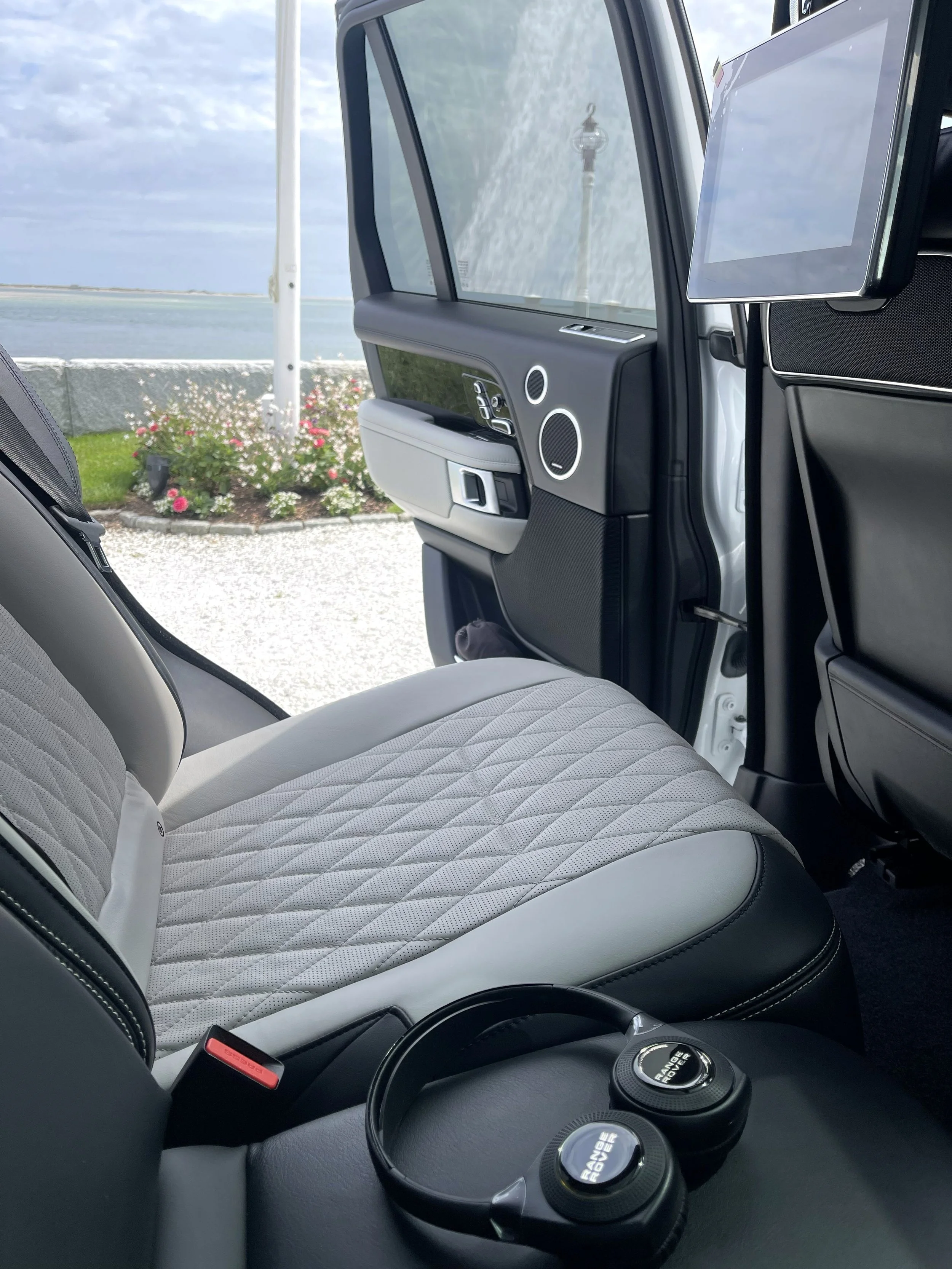 Inside a vehicle, showing a quilted car seat, dashboard, and a large monitor, with a view of flowers, a stone wall, and the ocean outside the window.