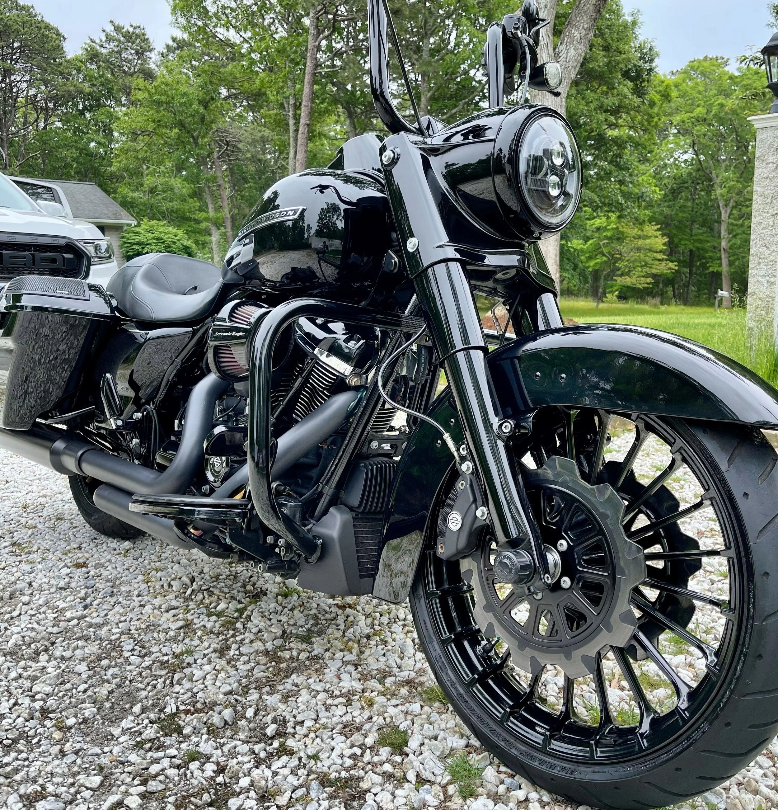 Close-up of a black Harley-Davidson motorcycle parked on a gravel driveway with green trees and a house in the background.