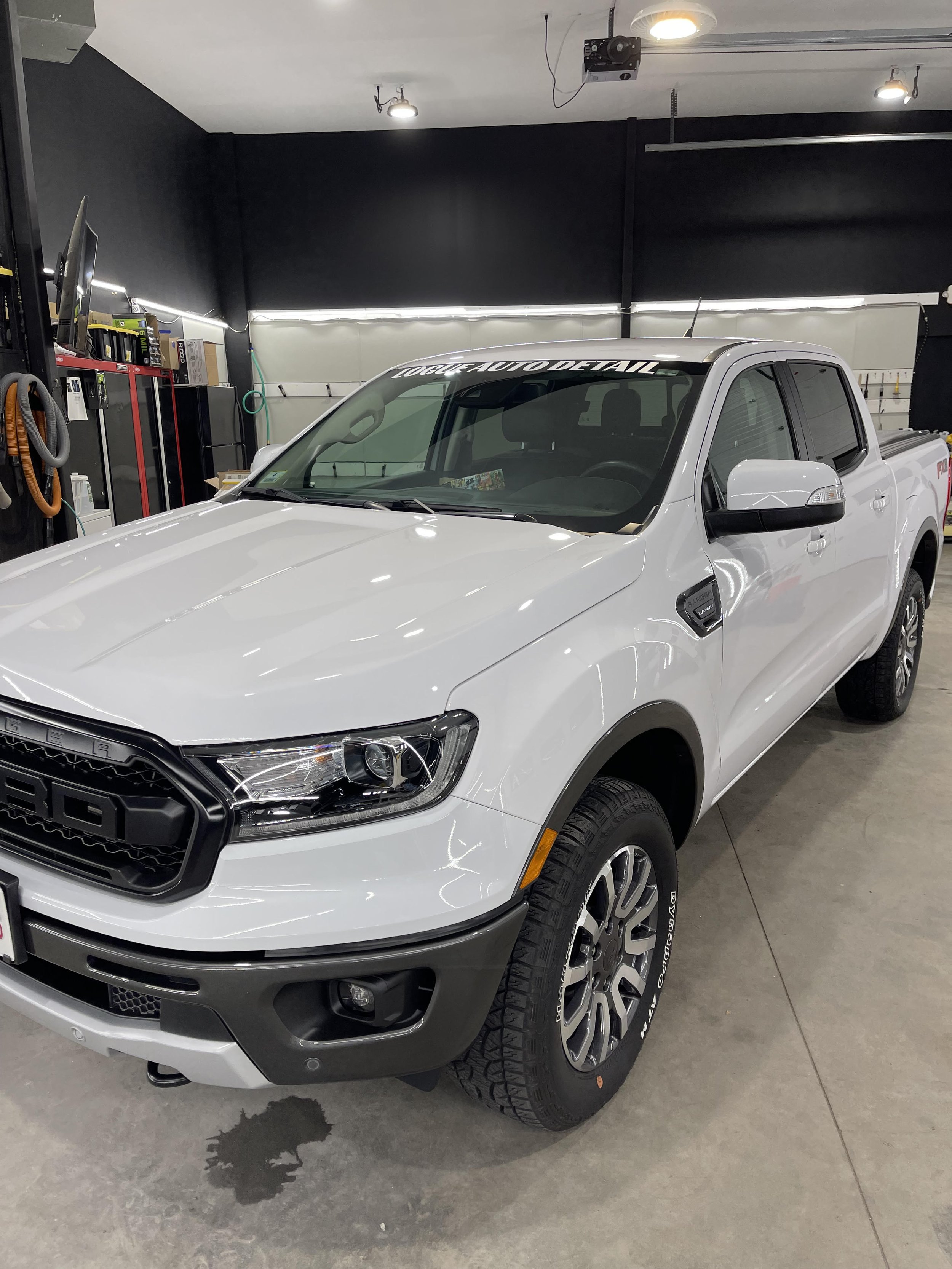 A white pickup truck parked inside a garage or workshop, with tools and equipment along the walls and a wet stain on the concrete floor.