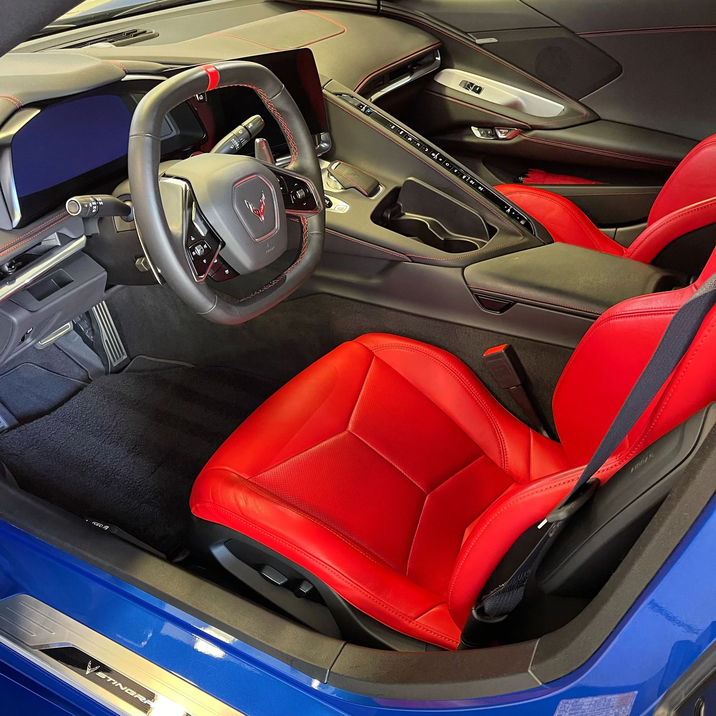 The interior of a sports car featuring a black dashboard with red stitching, a steering wheel with a Corvette logo, and red leather seats.