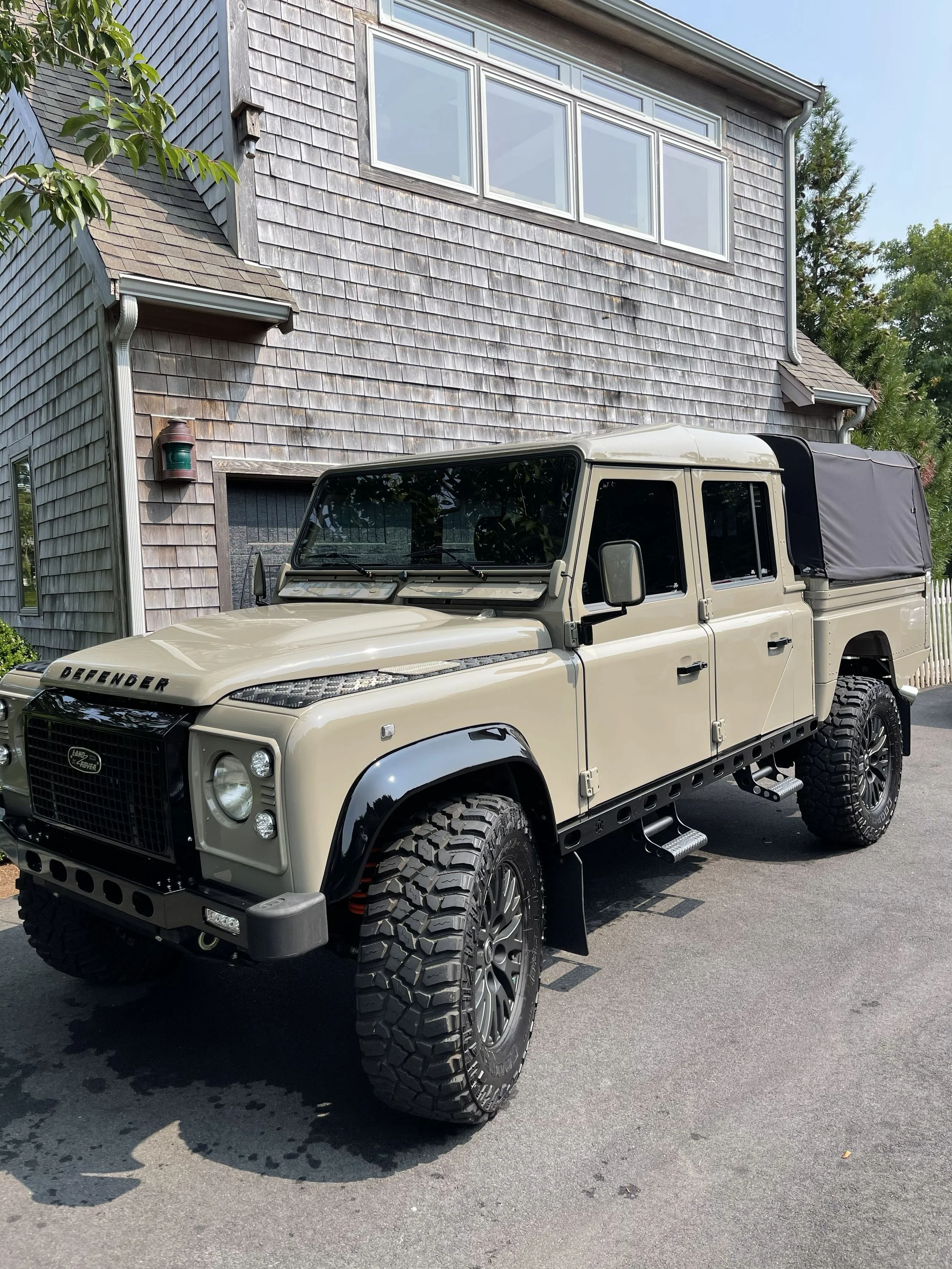 A beige Land Rover Defender parked in front of a gray wooden house with shingle siding. The house has a large window on the upper level and a smaller window on the lower level. There is a green tree on the left and taller evergreen trees on the right
