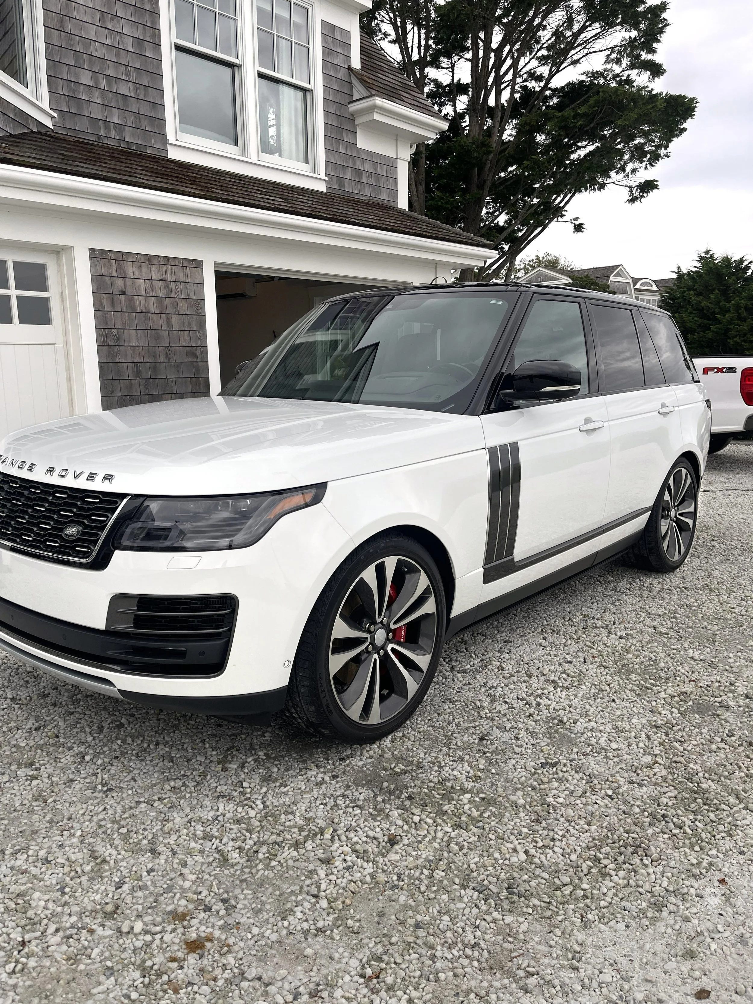 A white Range Rover SUV parked on a gravel driveway in front of a house with grey siding, white trim, and multiple windows. A tree is visible in the background along with part of a white truck.