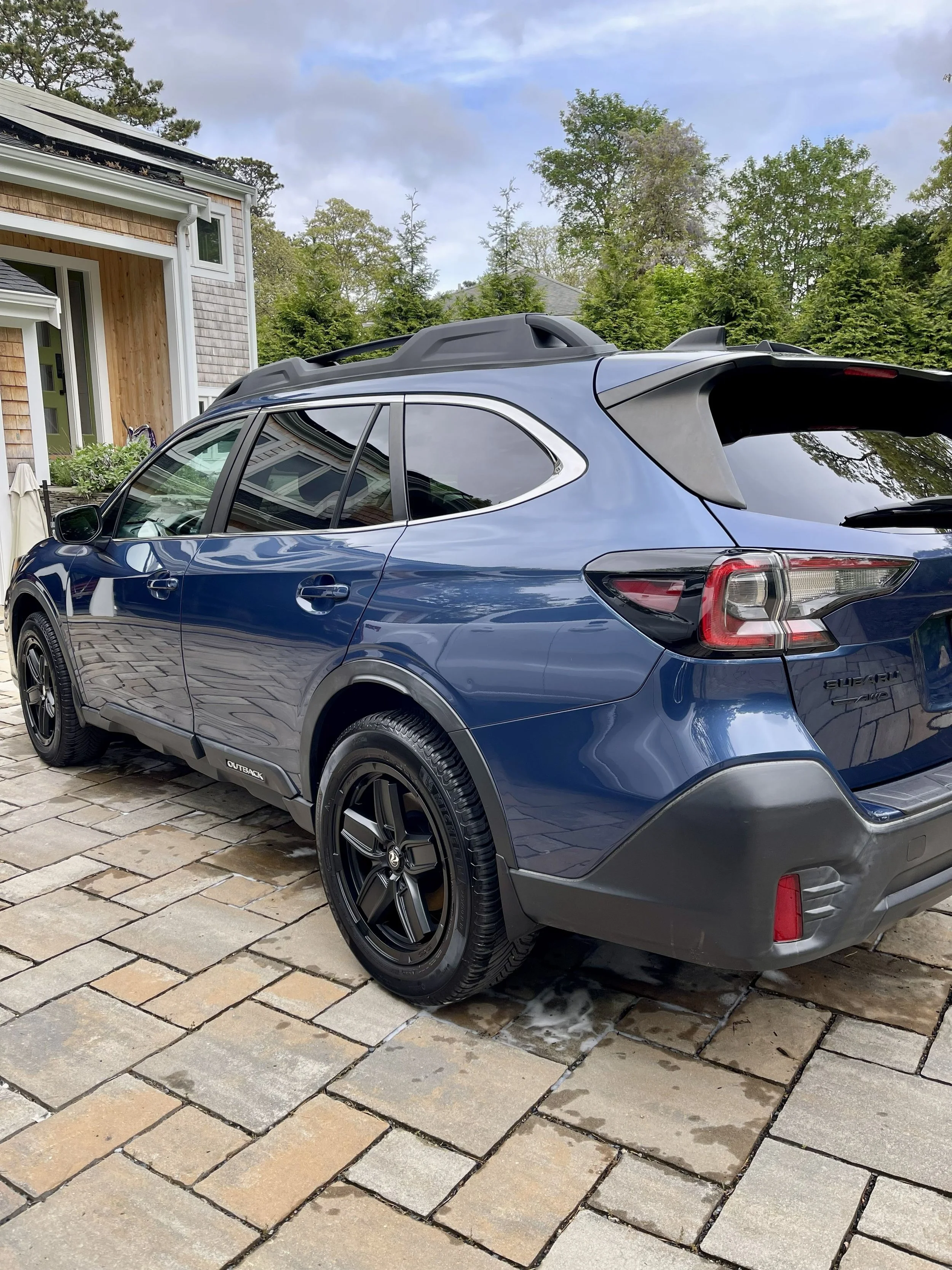 A blue Subaru SUV parked on a stone driveway in front of a house with wooden and shingled siding, surrounded by green trees, with a cloudy sky above.