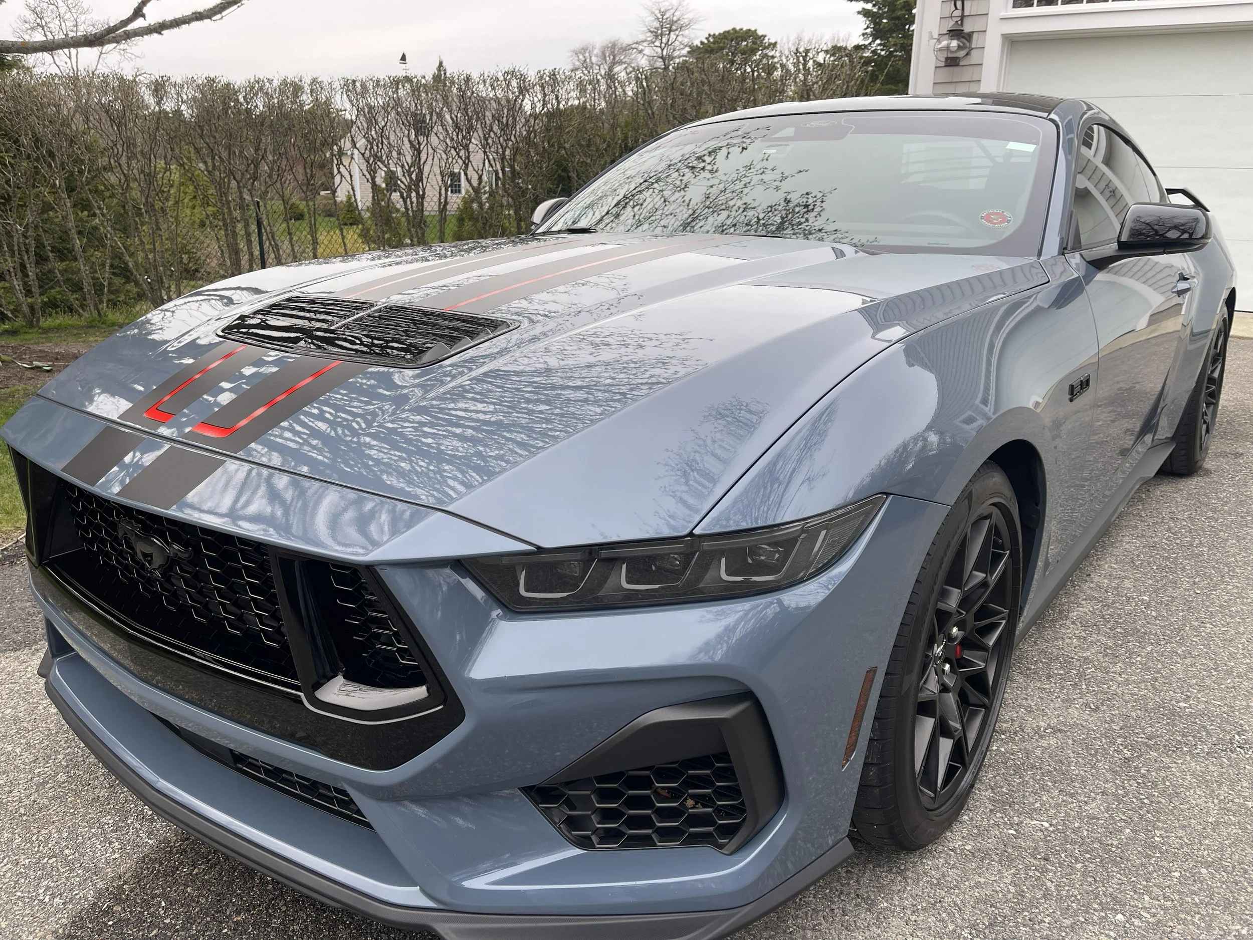 A gray sports car with black and red racing stripes parked on a driveway outside a house, with trees and a yard in the background.