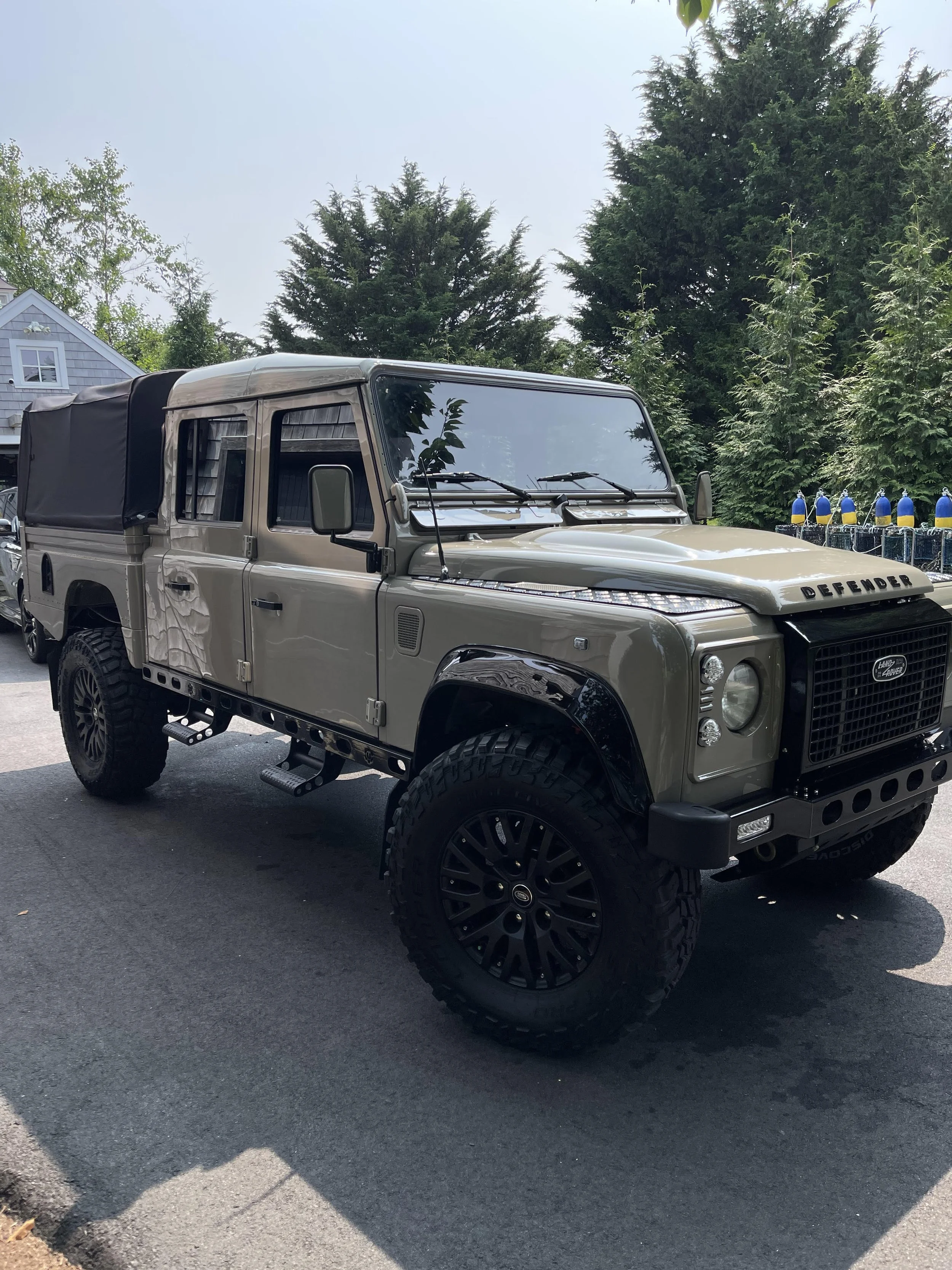 A beige Land Rover Defender parked outdoors with a black roof cover, large off-road tires, and black rims, with lush green trees in the background.