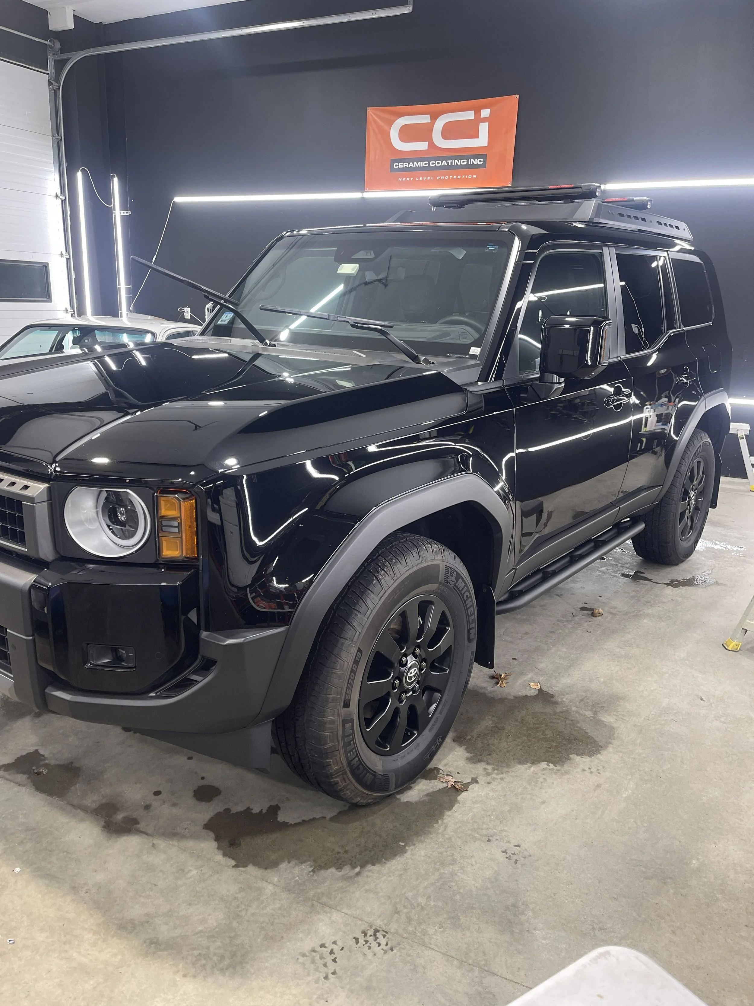 Black SUV parked inside a garage with a blue wall, displaying the Ceramic Coating Inc banner, a car with distinctive headlights, and some leaves on the concrete floor.