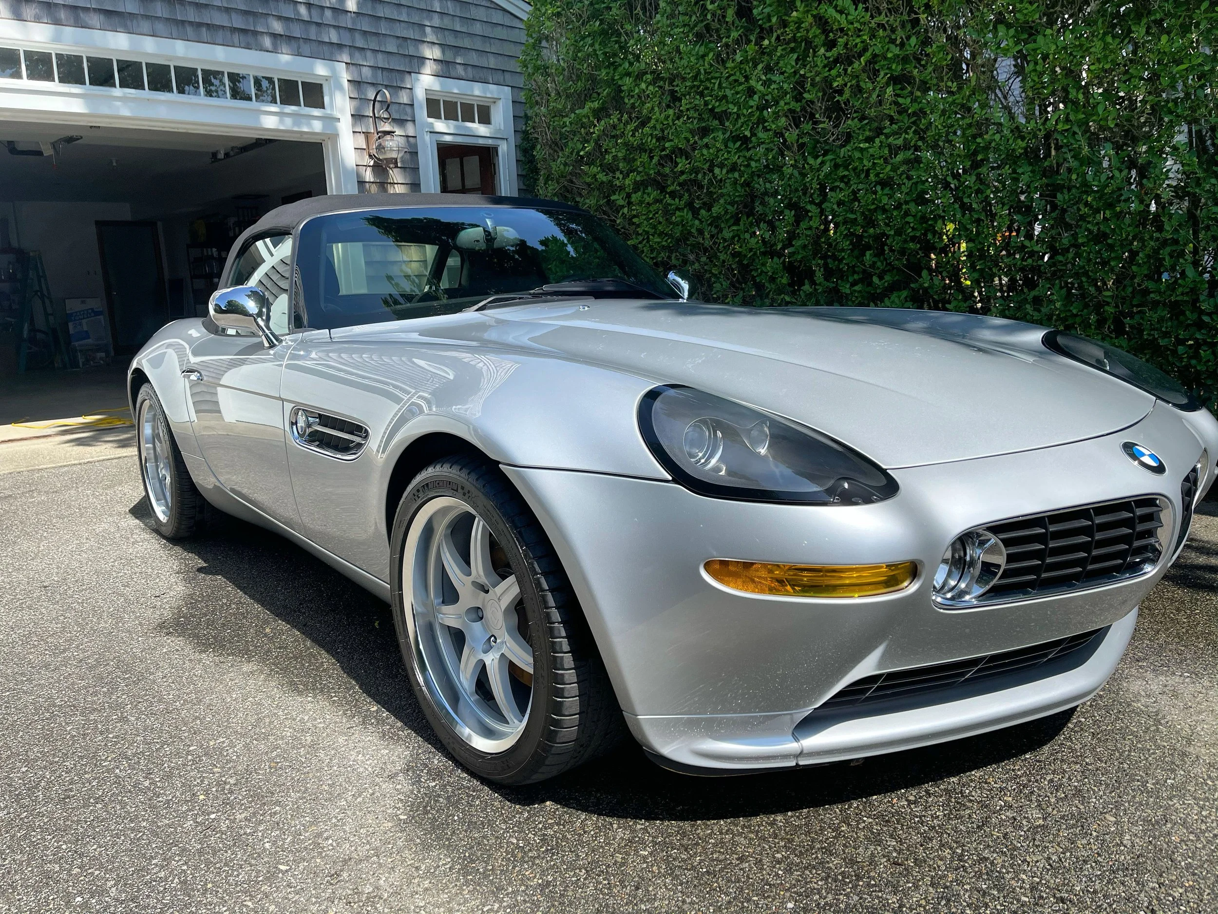 A silver BMW Z8 roadster convertible car parked in a driveway in front of a garage, with a green hedge on the right side.
