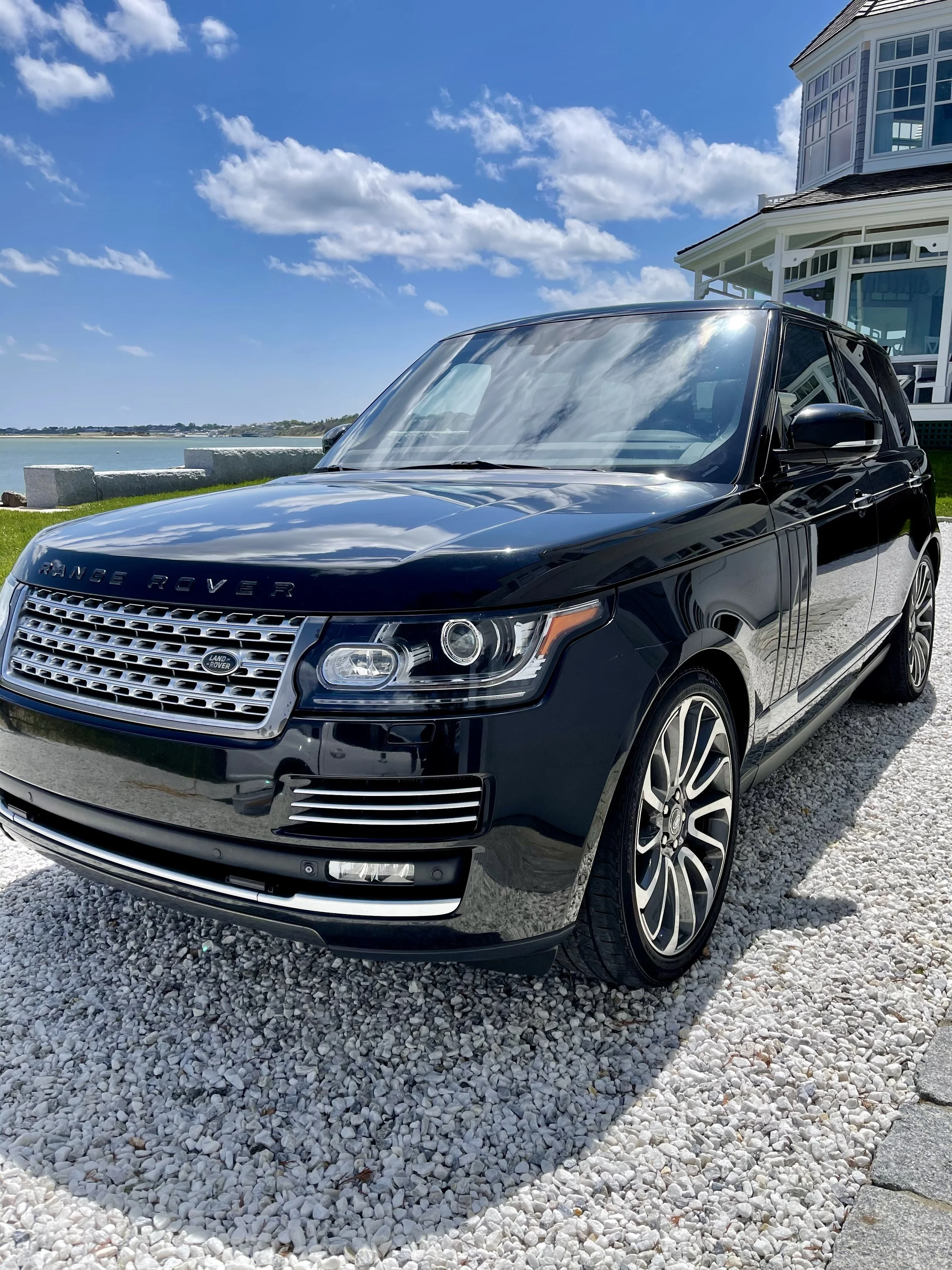 Black Range Rover parked on white gravel near a waterfront with a house in the background and partly cloudy sky.