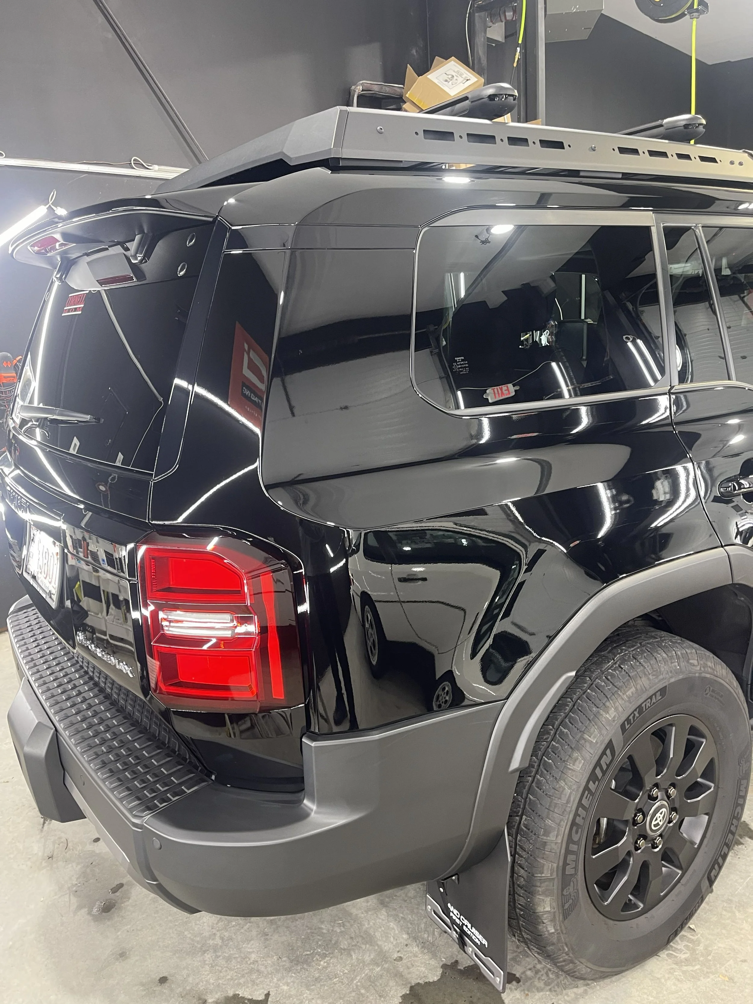 Black mid-size SUV parked indoors, showing the rear and side of the vehicle with a visible tire, taillight, and rear window. The SUV has a roof rack and a two-tone color scheme with gray accents.