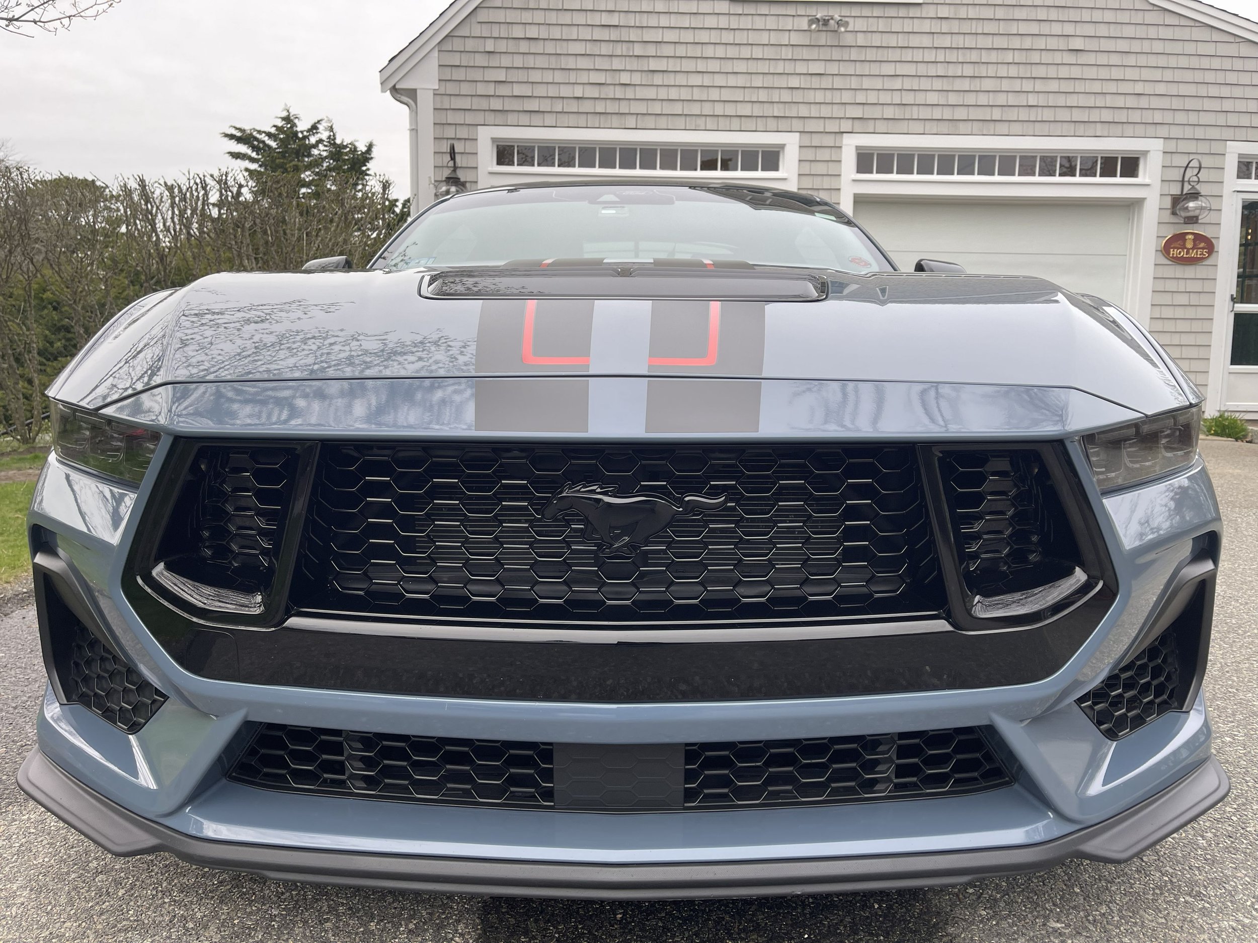 Front view of a gray Ford Mustang sports car with black racing stripes parked in front of a house.