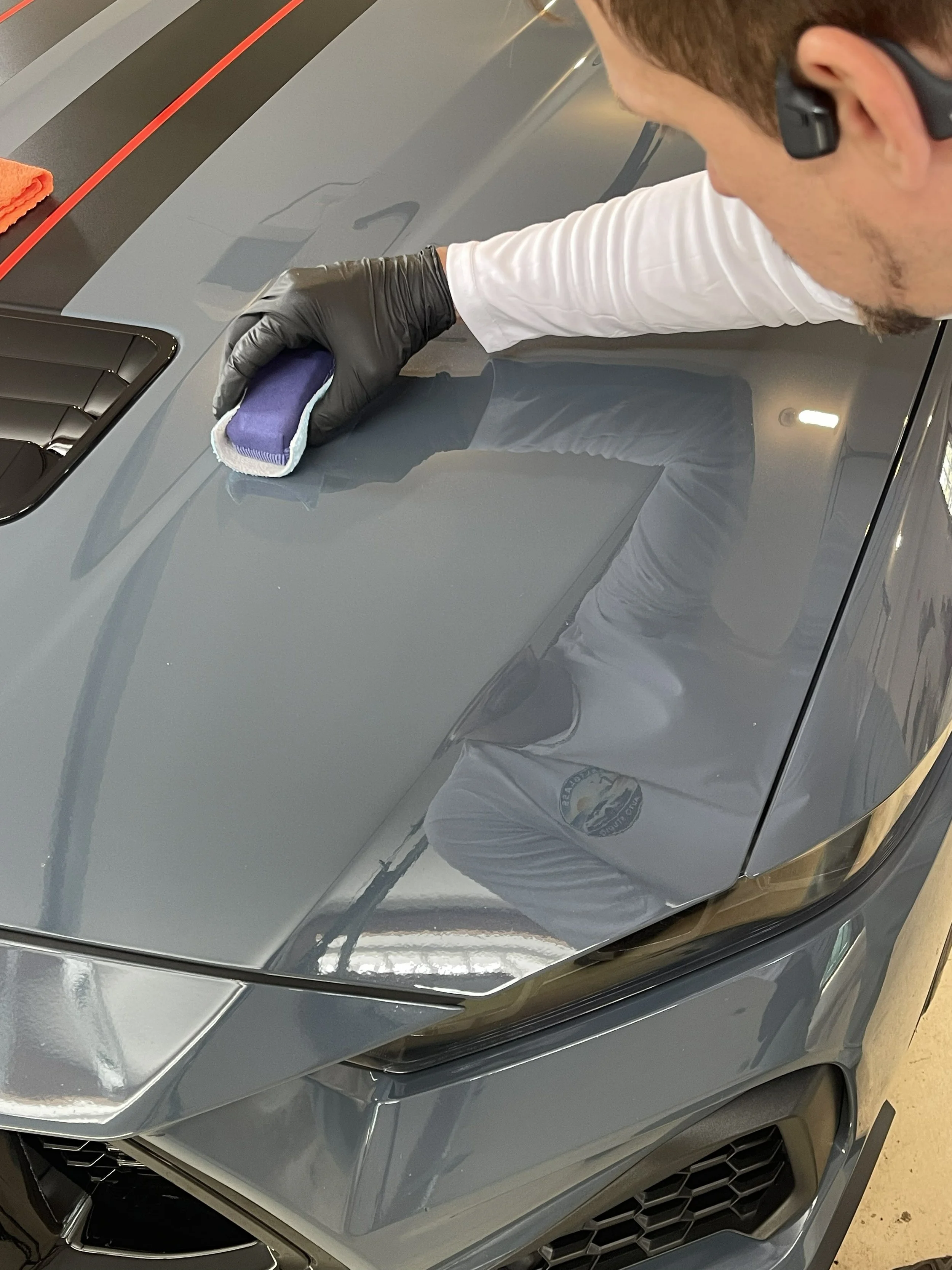 A person wearing black gloves and a microphone headset is cleaning the hood of a gray car with a sponge or cloth.