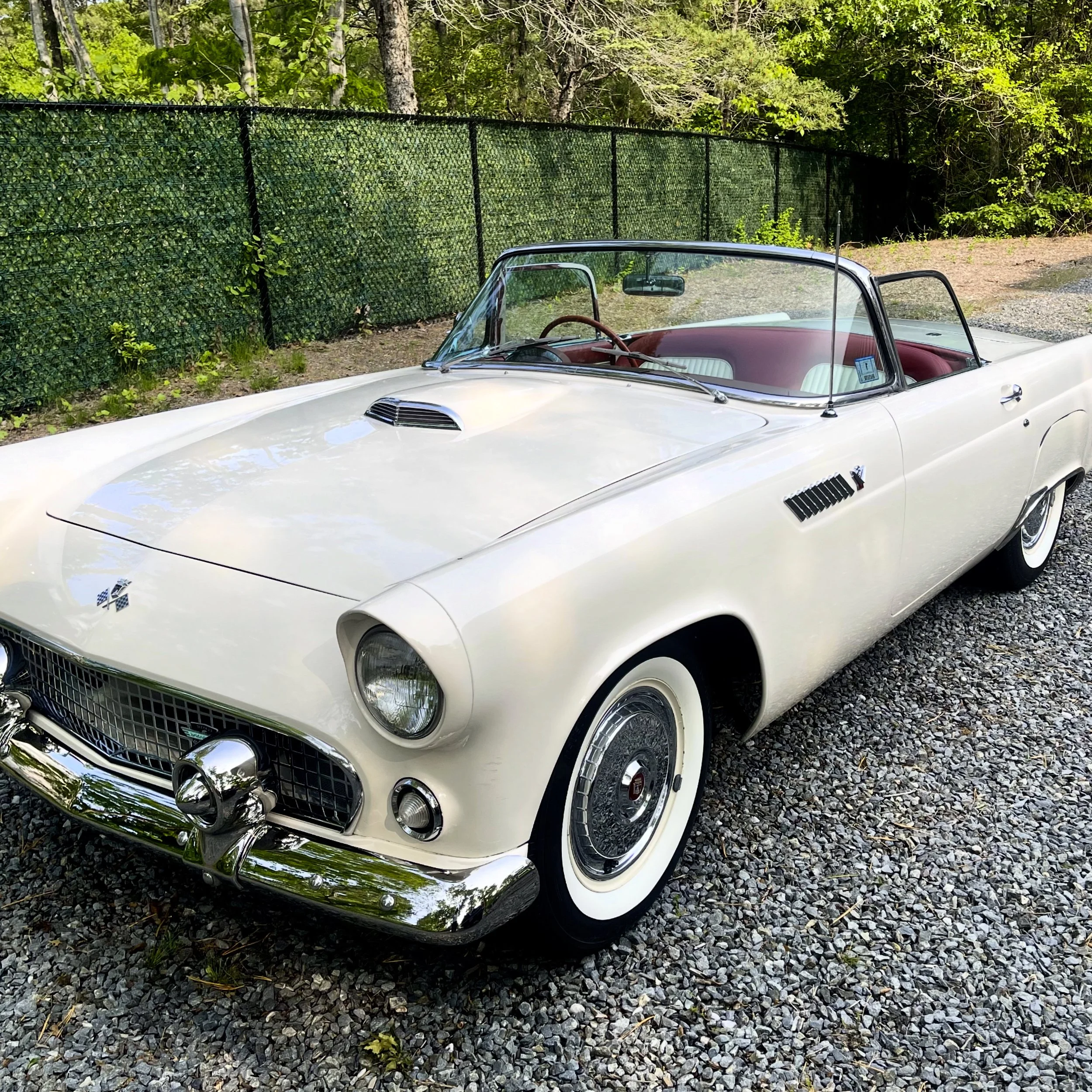 A vintage white convertible car parked on a gravel surface with a green netted fence and trees in the background.