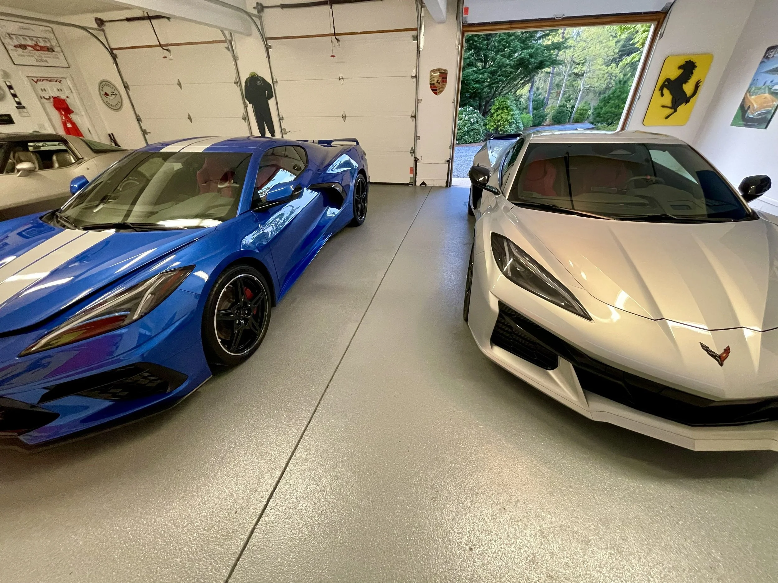 Inside a garage with two sports cars parked side by side, one blue with white racing stripes and one silver, with a view of greenery outside through the open garage door, and various car posters and memorabilia on the walls.
