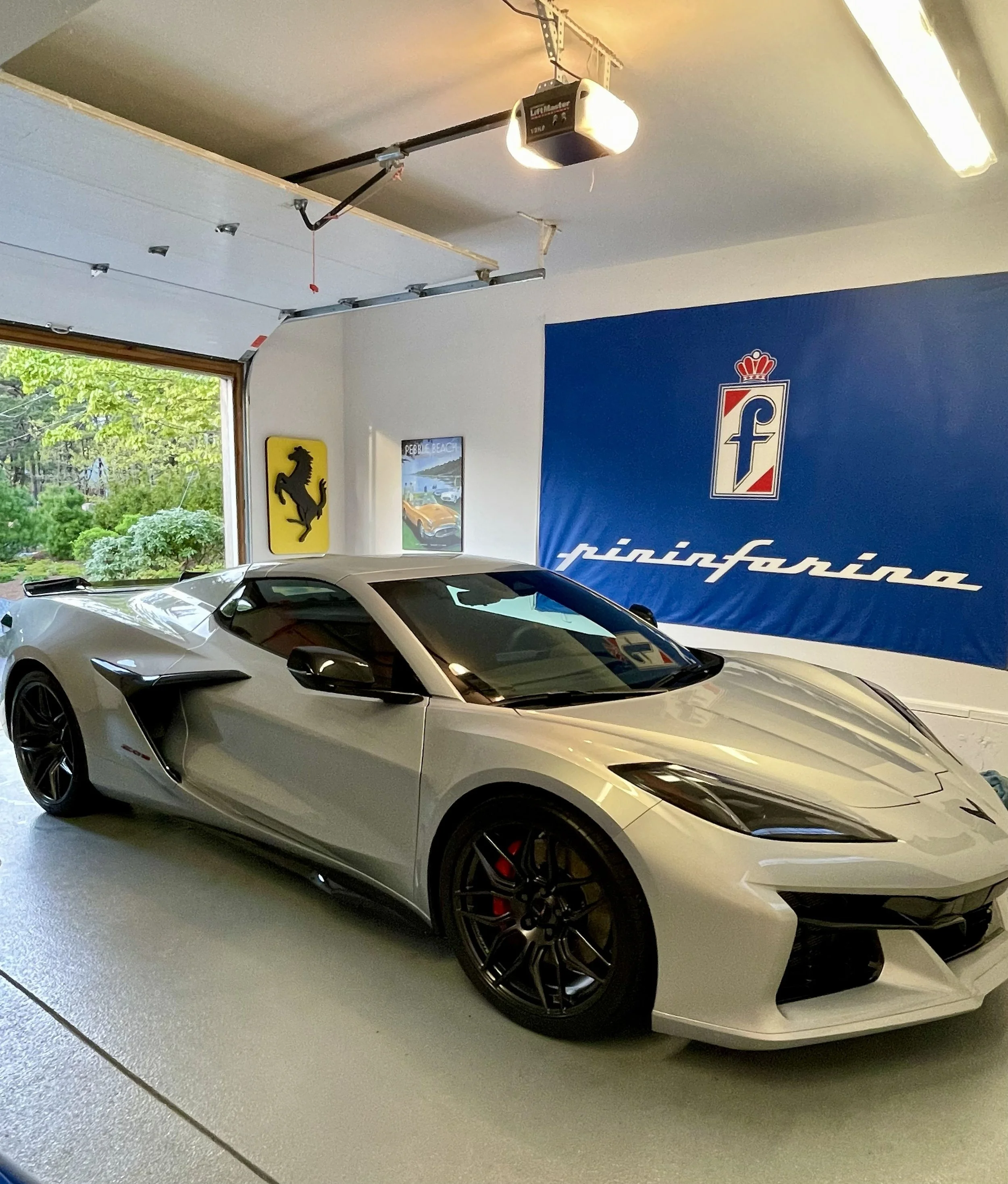 Silver sports car with black wheels parked inside a garage, with a large Ferrari logo wall decal and a blue banner behind it.
