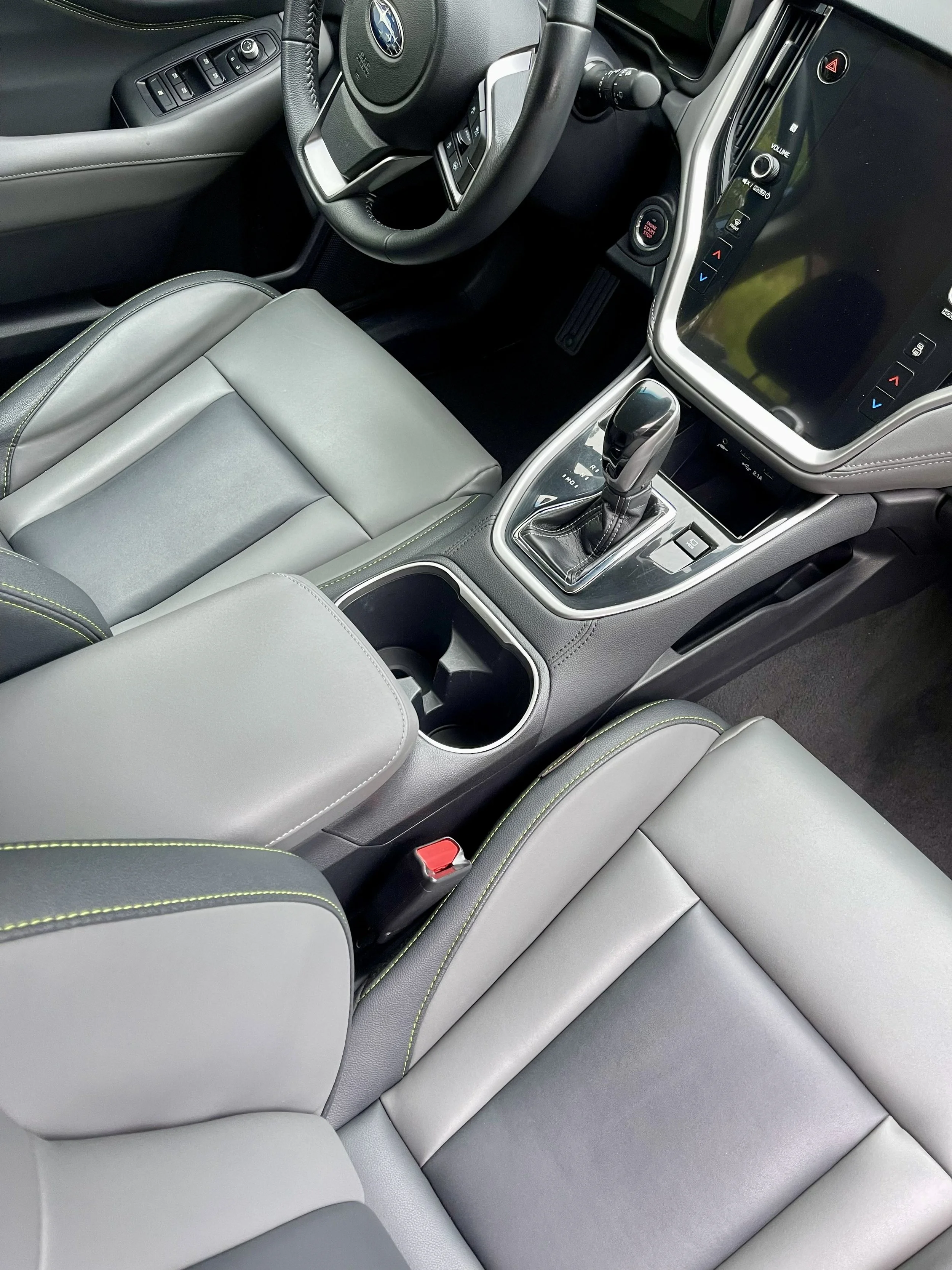 The interior of a modern vehicle showing the front passenger seat, center console with gear shift, part of the dashboard, and steering wheel with controls. The seats are light gray with green stitching accents.