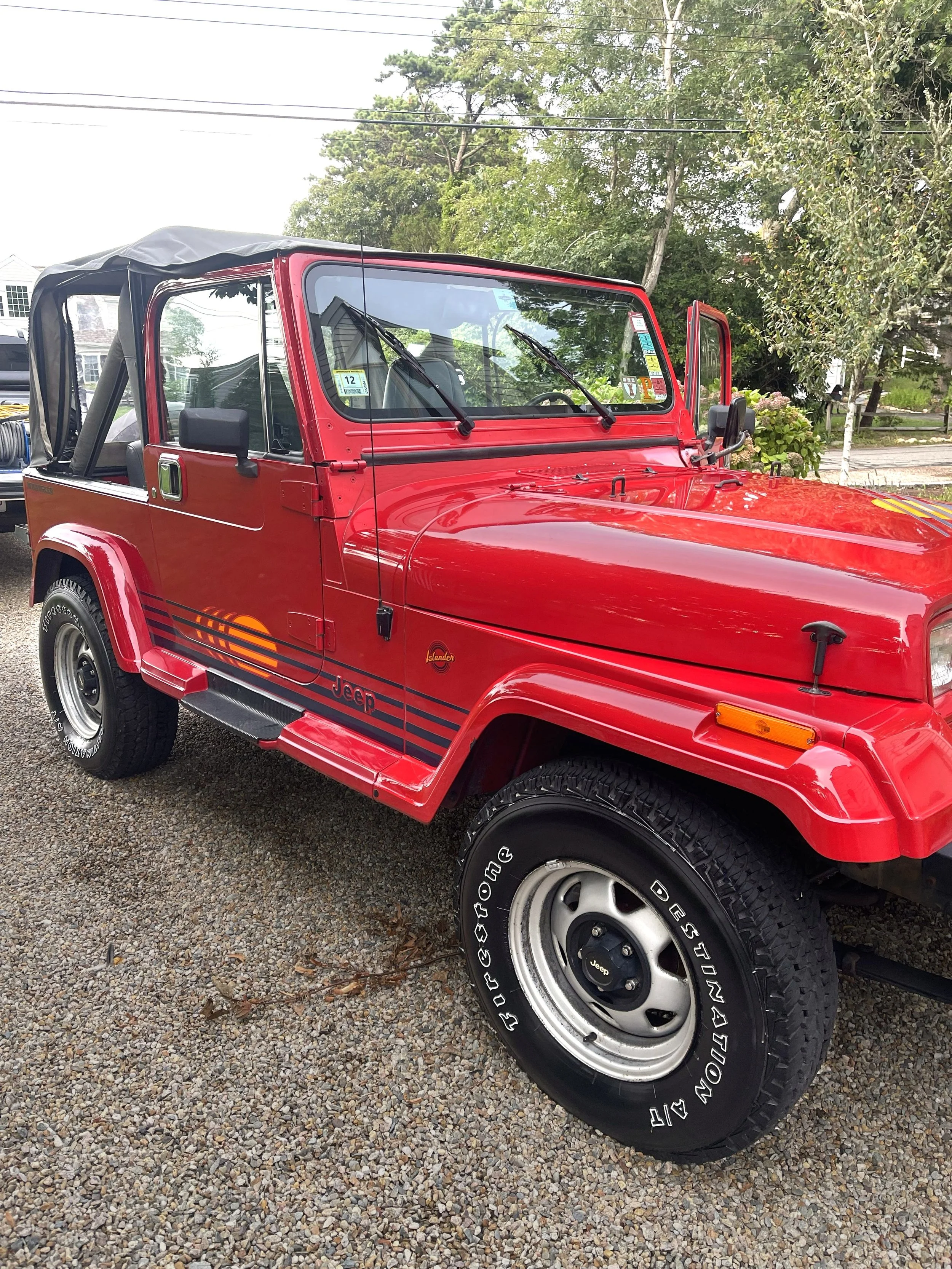 A red Jeep with black and orange stripe decals, parked on a gravel driveway. The top of the Jeep is black and partially open, with a rearview mirror on the passenger side visible. The background has trees, a house, and some greenery.