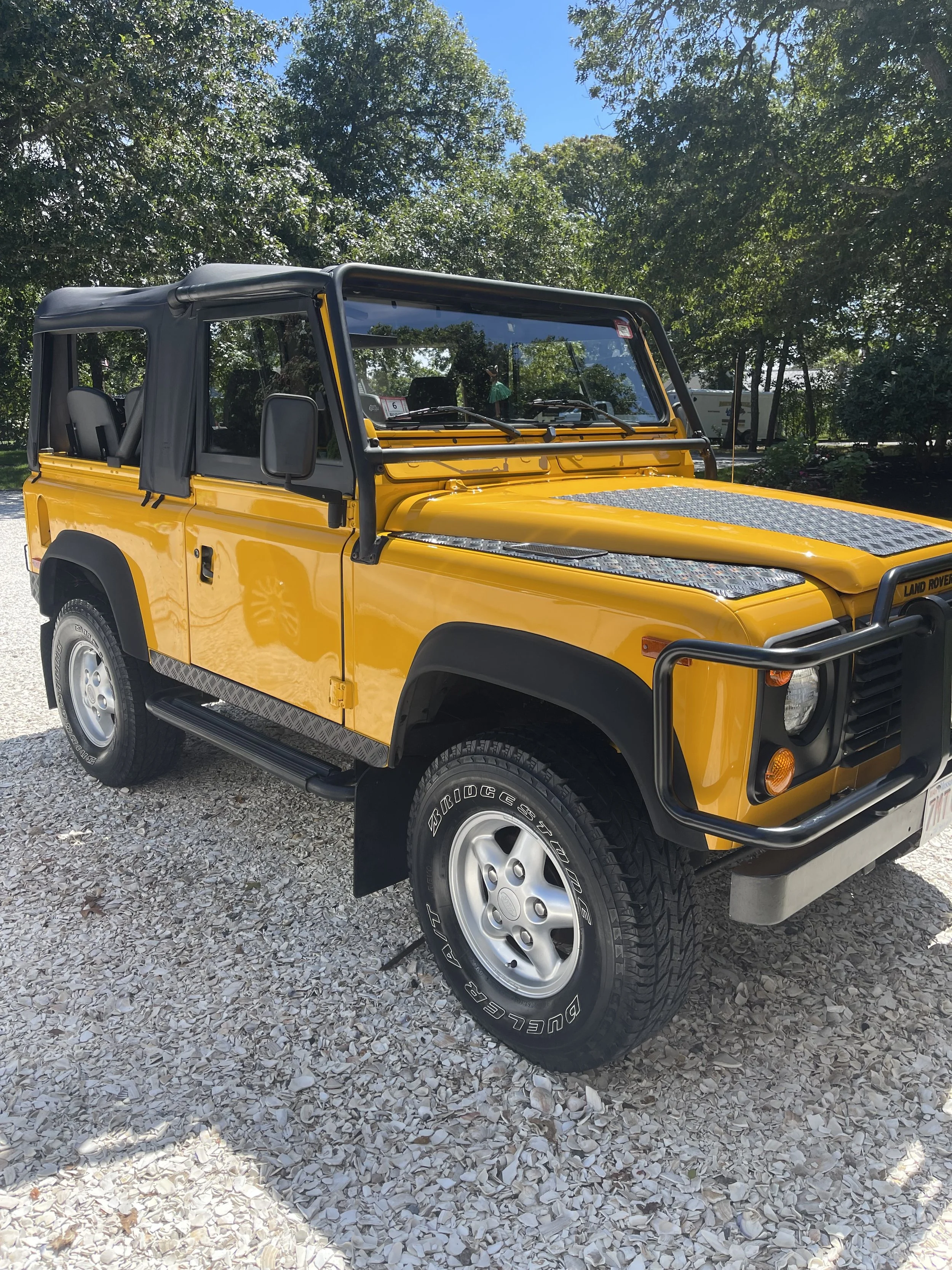 Yellow Land Rover Defender with a black soft top parked on gravel, surrounded by green trees.