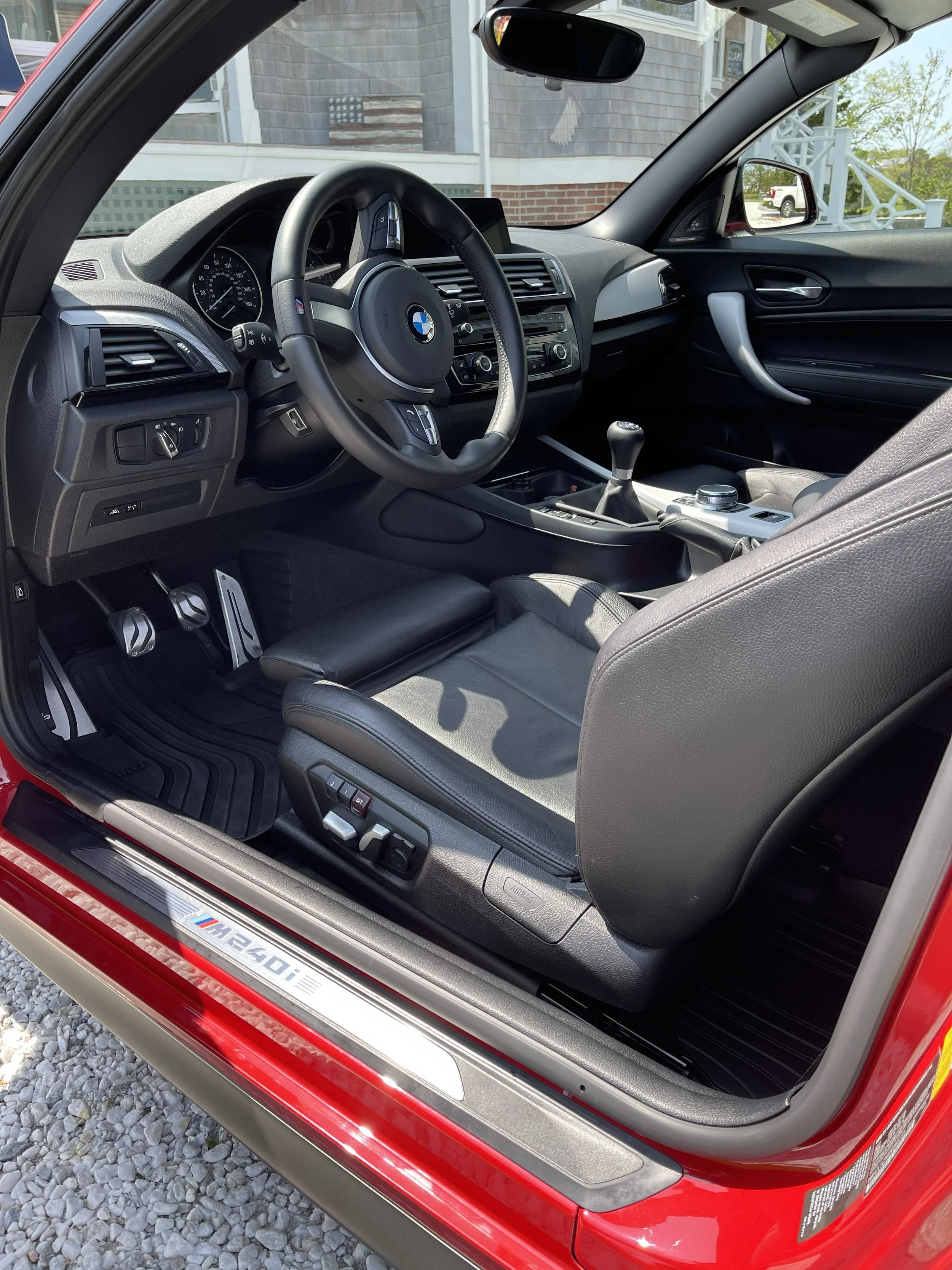 Interior of a red BMW M4, showing the steering wheel, gear shift, dashboard, and black leather seats, parked outside a house with gravel driveway.