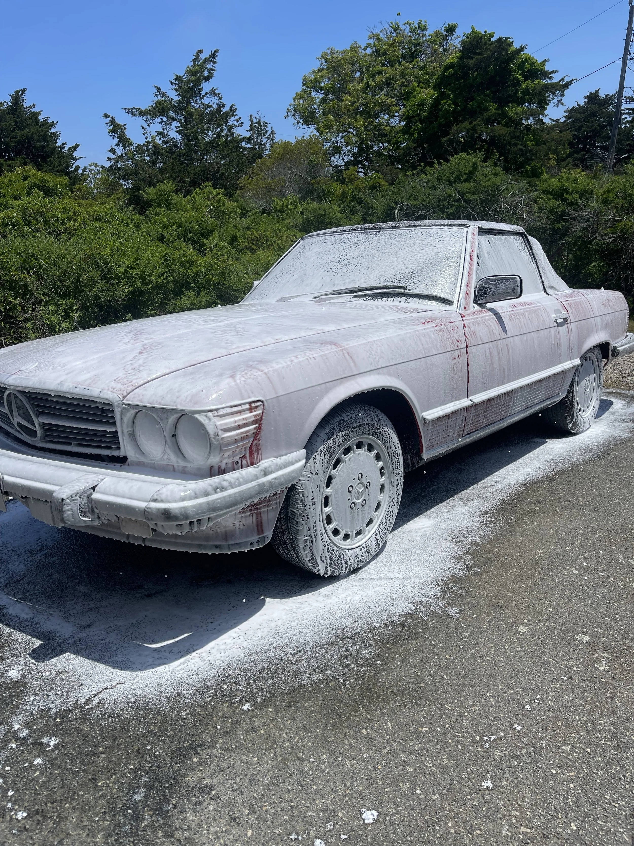 A white vintage Mercedes-Benz car covered in foam and soap, parked on a asphalt road with greenery and trees in the background under a clear blue sky.