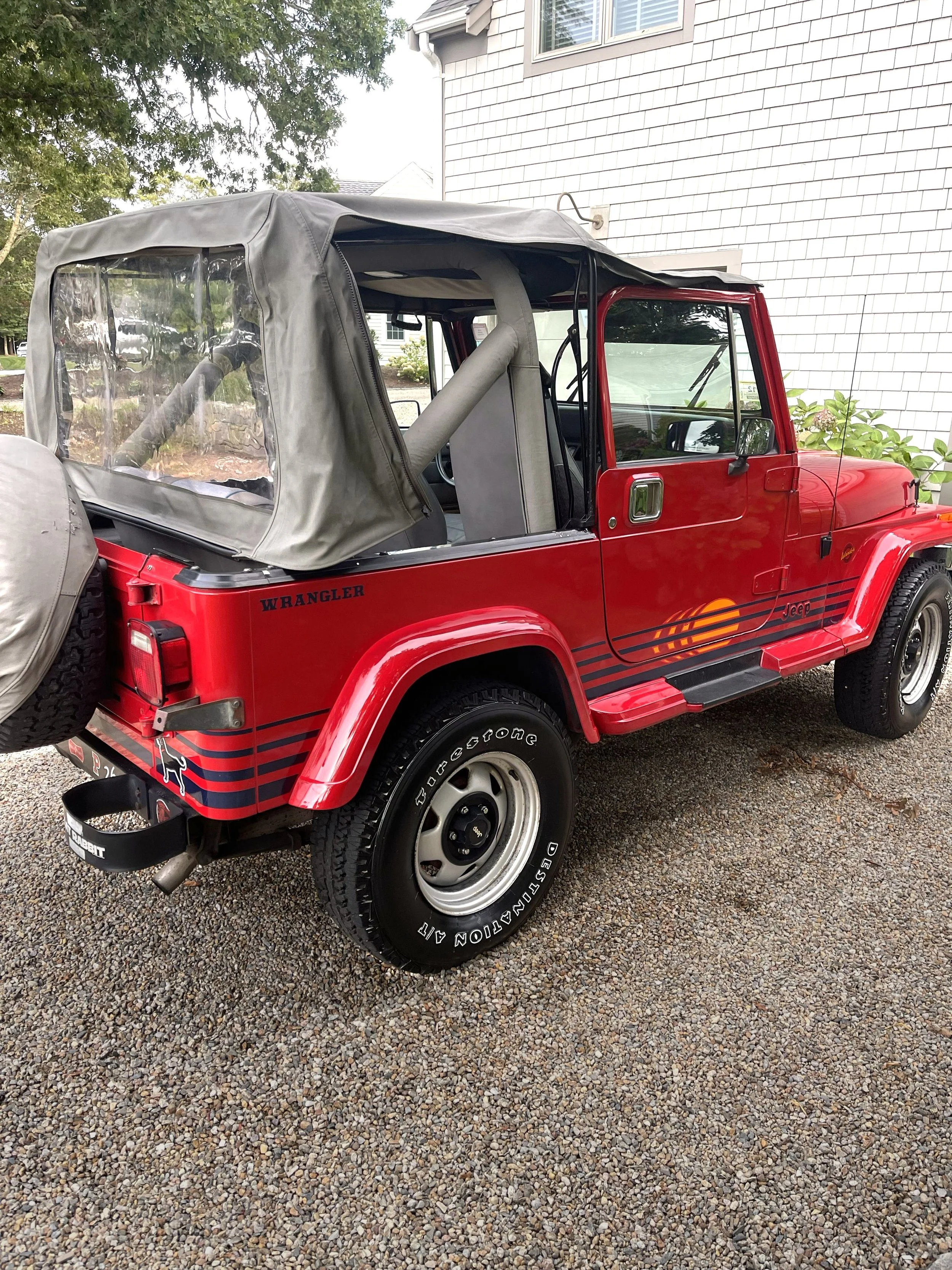 A red Jeep Wrangler SUV with a gray soft top, parked on a gravel driveway next to a white house with gray shingles and greenery.