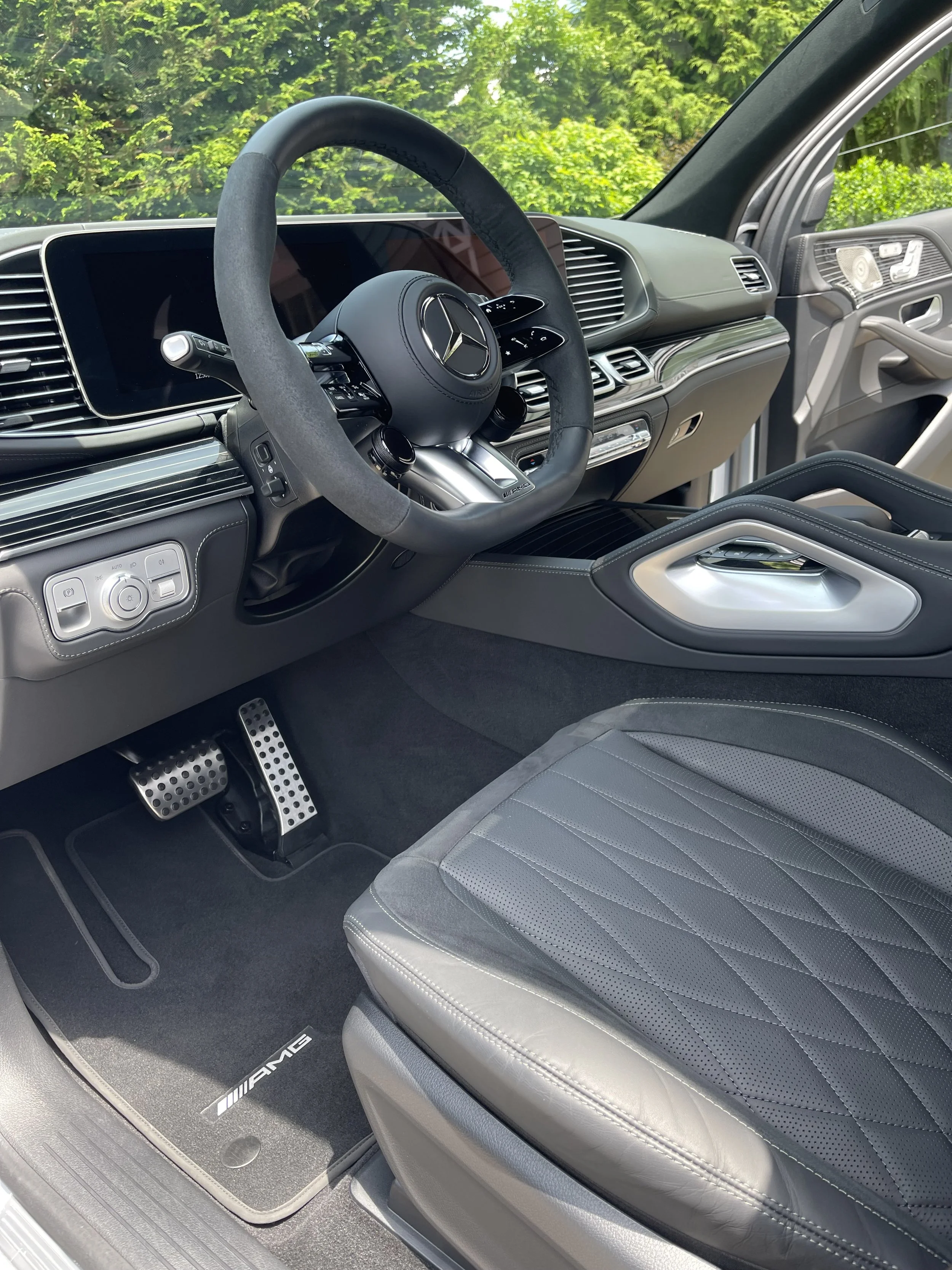 Interior of a luxury Mercedes-Benz AMG vehicle, showing a black leather seat with silver stitching, a steering wheel with Mercedes-Benz emblem, digital dashboard, and gear shift. The car's floor mat has AMG branding.