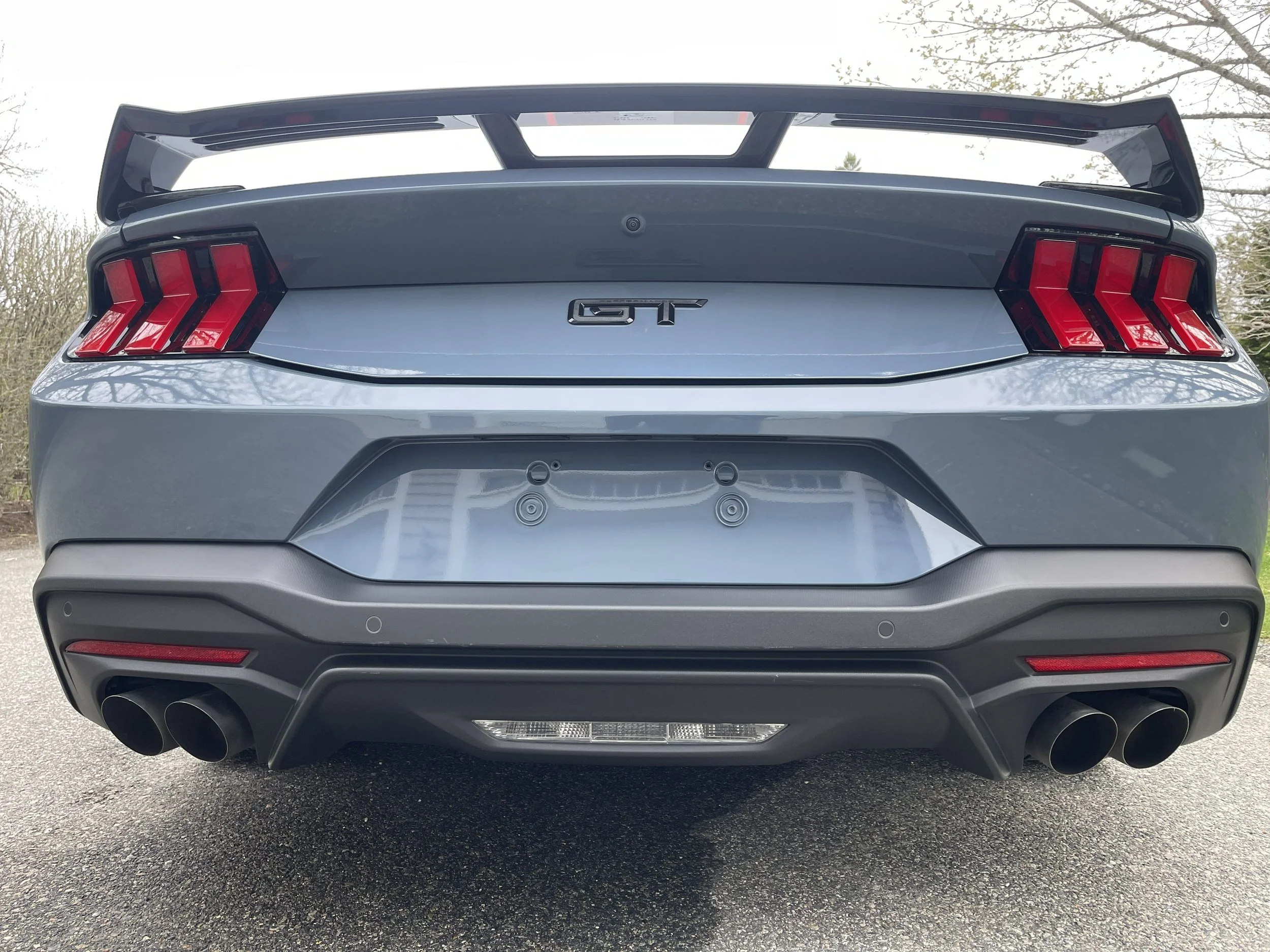 The rear view of a silver Ford Mustang GT with a large rear spoiler, dark taillights, dual exhaust pipes, and a visible GT badge.