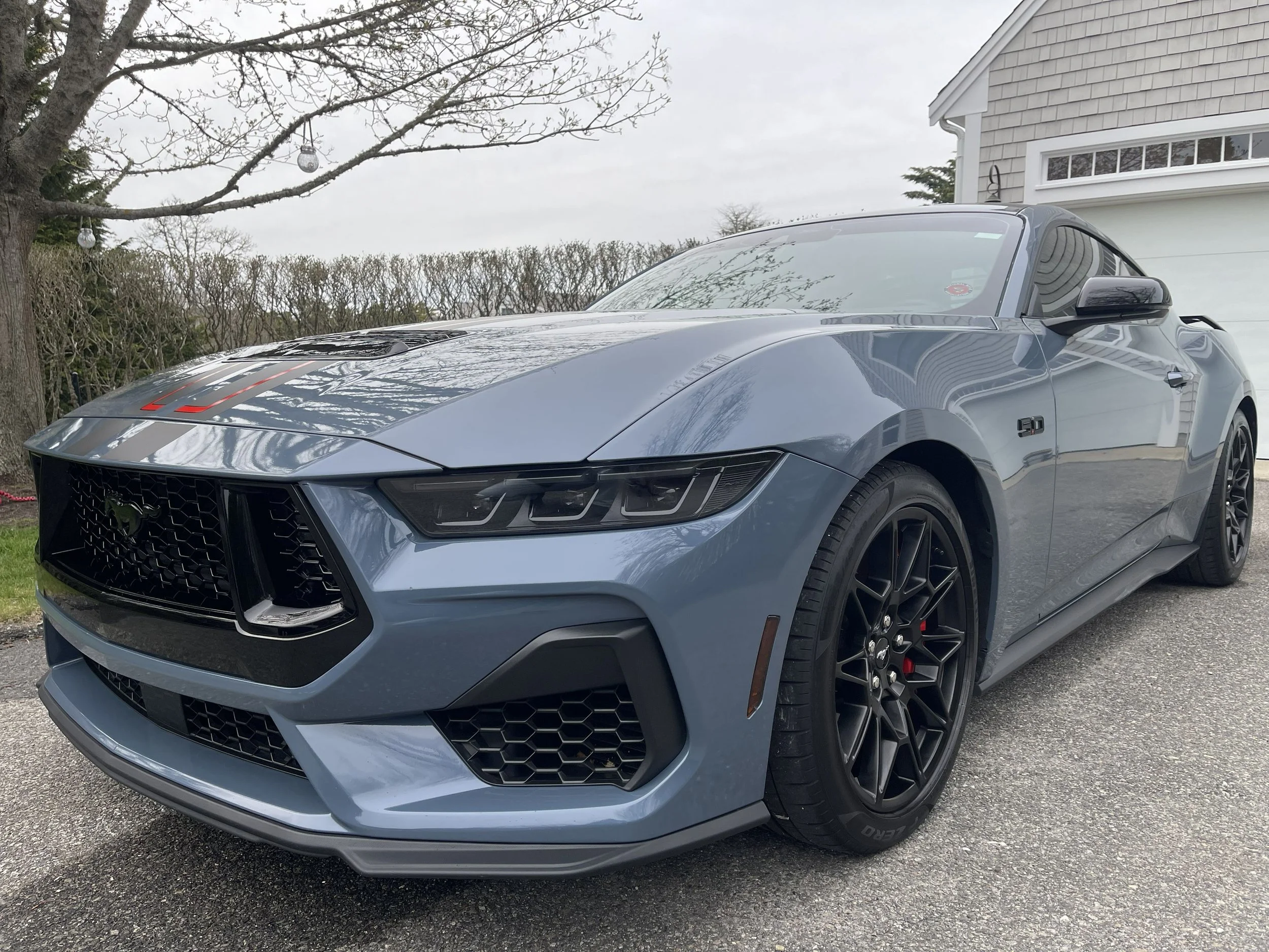 A gray Ford Mustang sports car parked on a driveway next to a house with gray shingles. A tree with bare branches is in the background, and there are hanging glass ornaments from the tree.