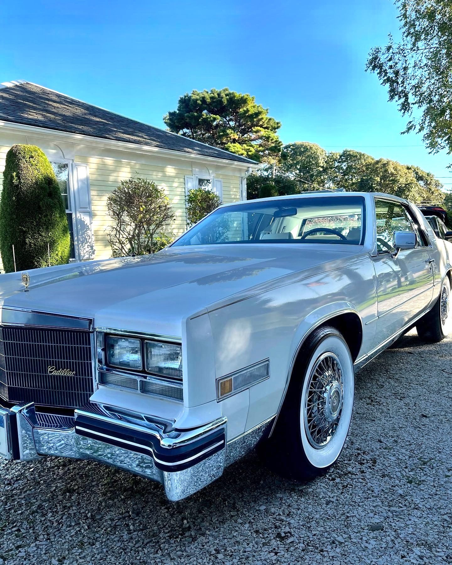 A vintage silver Cadillac car parked outdoors in front of a yellow house with white trim and a dark roof, surrounded by trees and bushes under a clear blue sky.