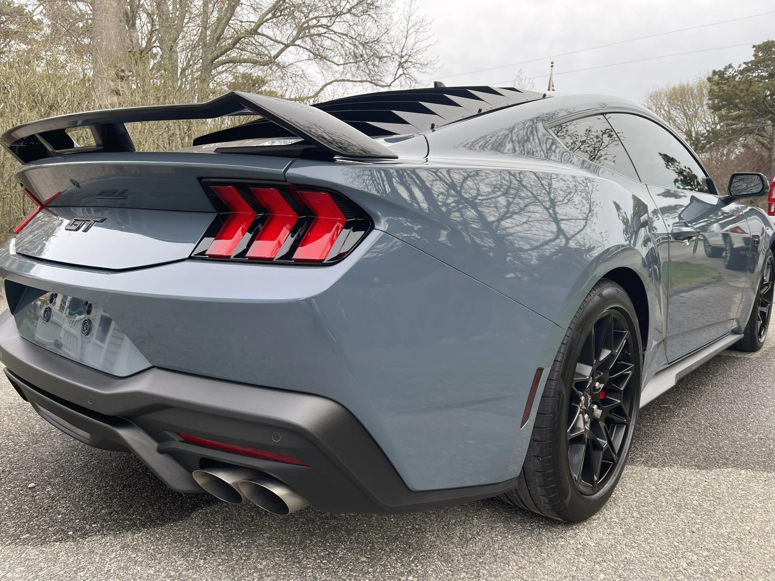A gray Ford Mustang GT sports car with a large rear spoiler, black wheels, and red taillights parked outdoors near trees.