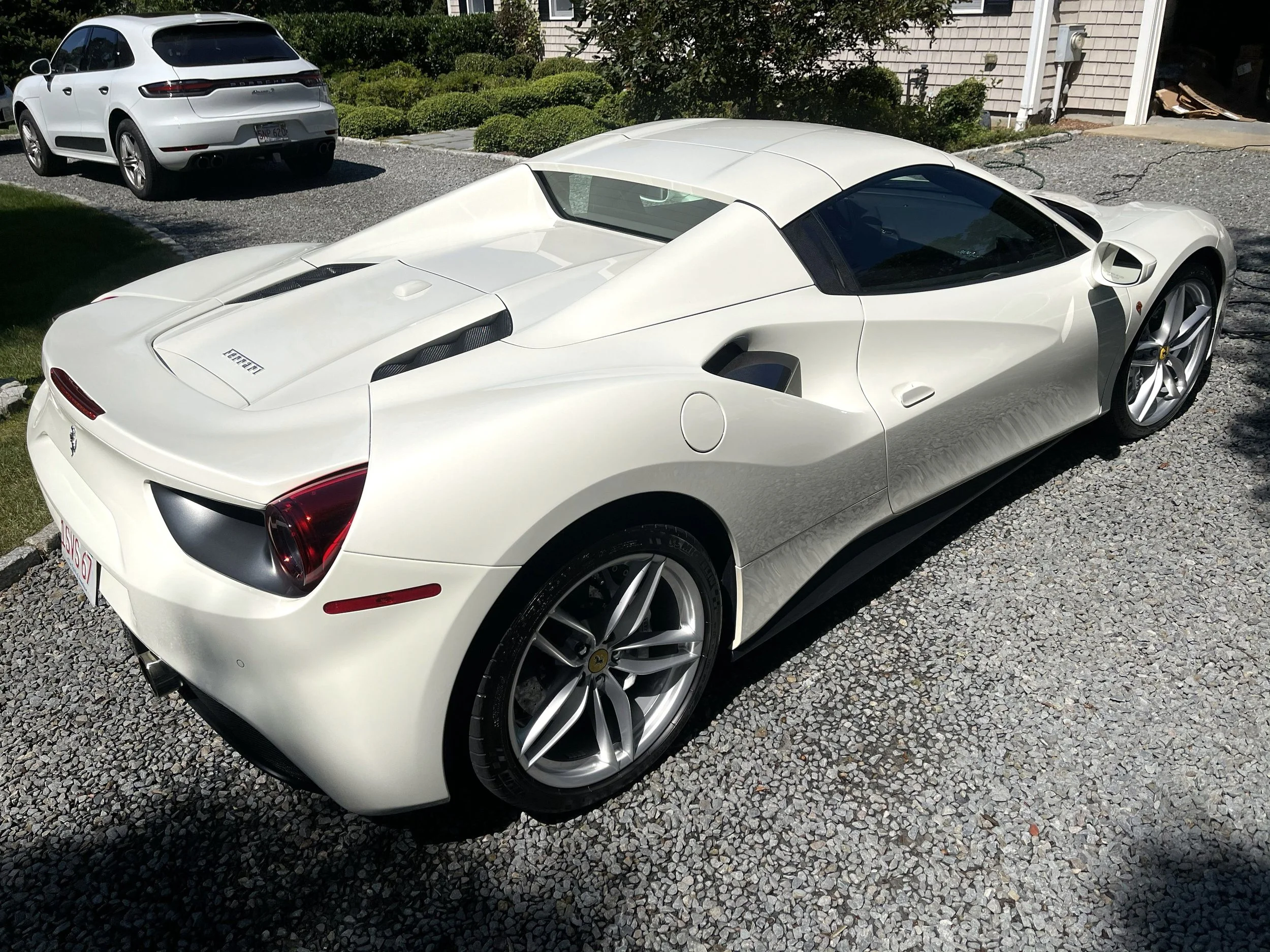 A white Ferrari sports car parked on a gravel driveway next to a house with a dodge SUV parked nearby. The Ferrari has a sleek design with black accents and large wheels.