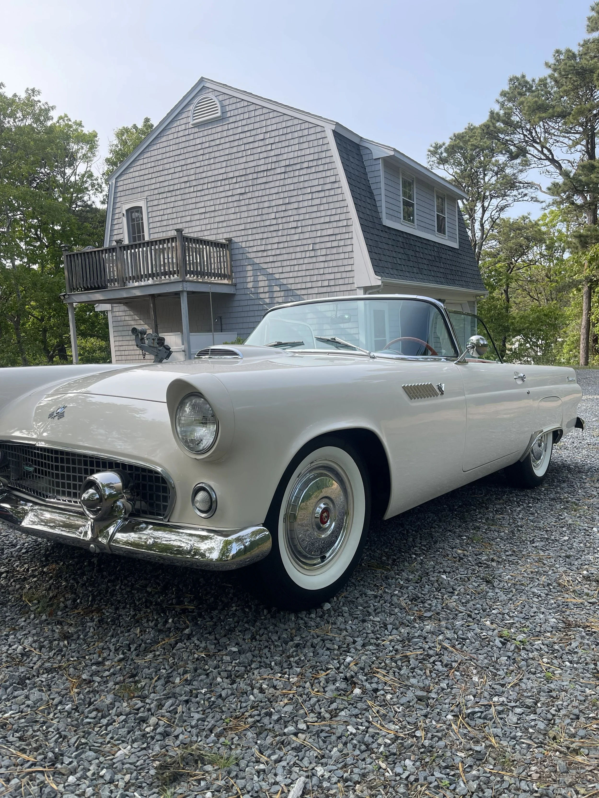 A white vintage Chevrolet Corvette convertible parked on a gravel driveway in front of a two-story house with gray shingles and black trim, surrounded by green trees.