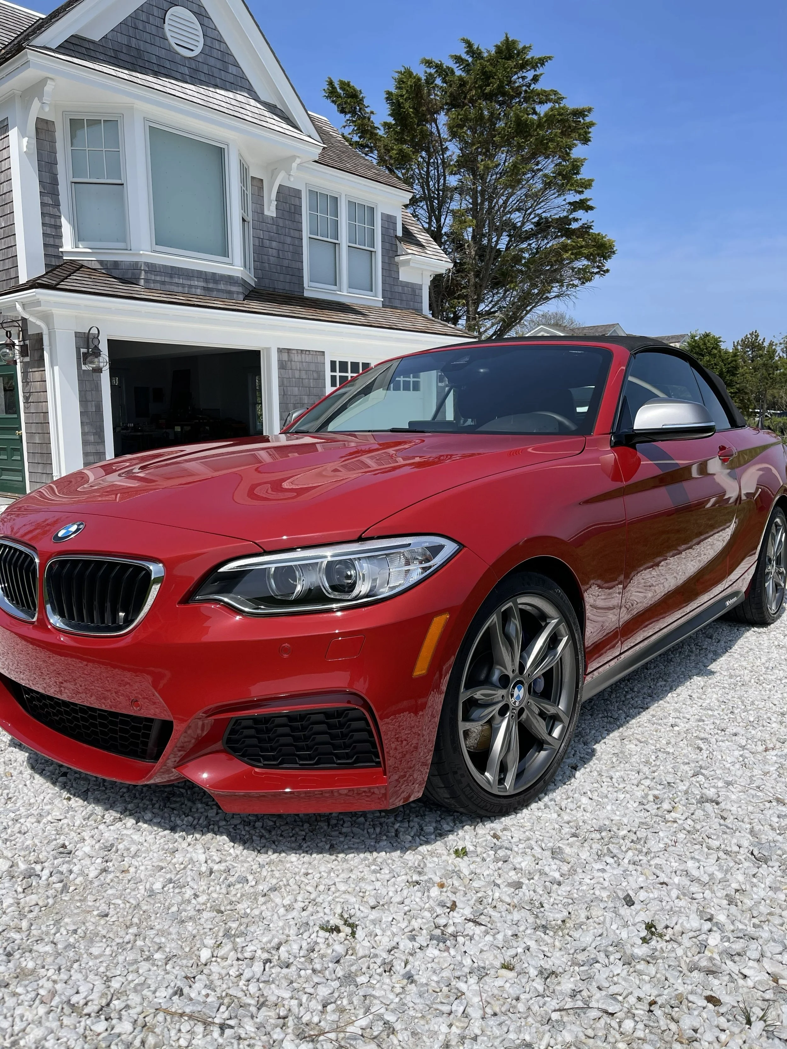Red BMW convertible parked on a gravel driveway in front of a gray house with white trim and large windows, with trees and a blue sky in the background.