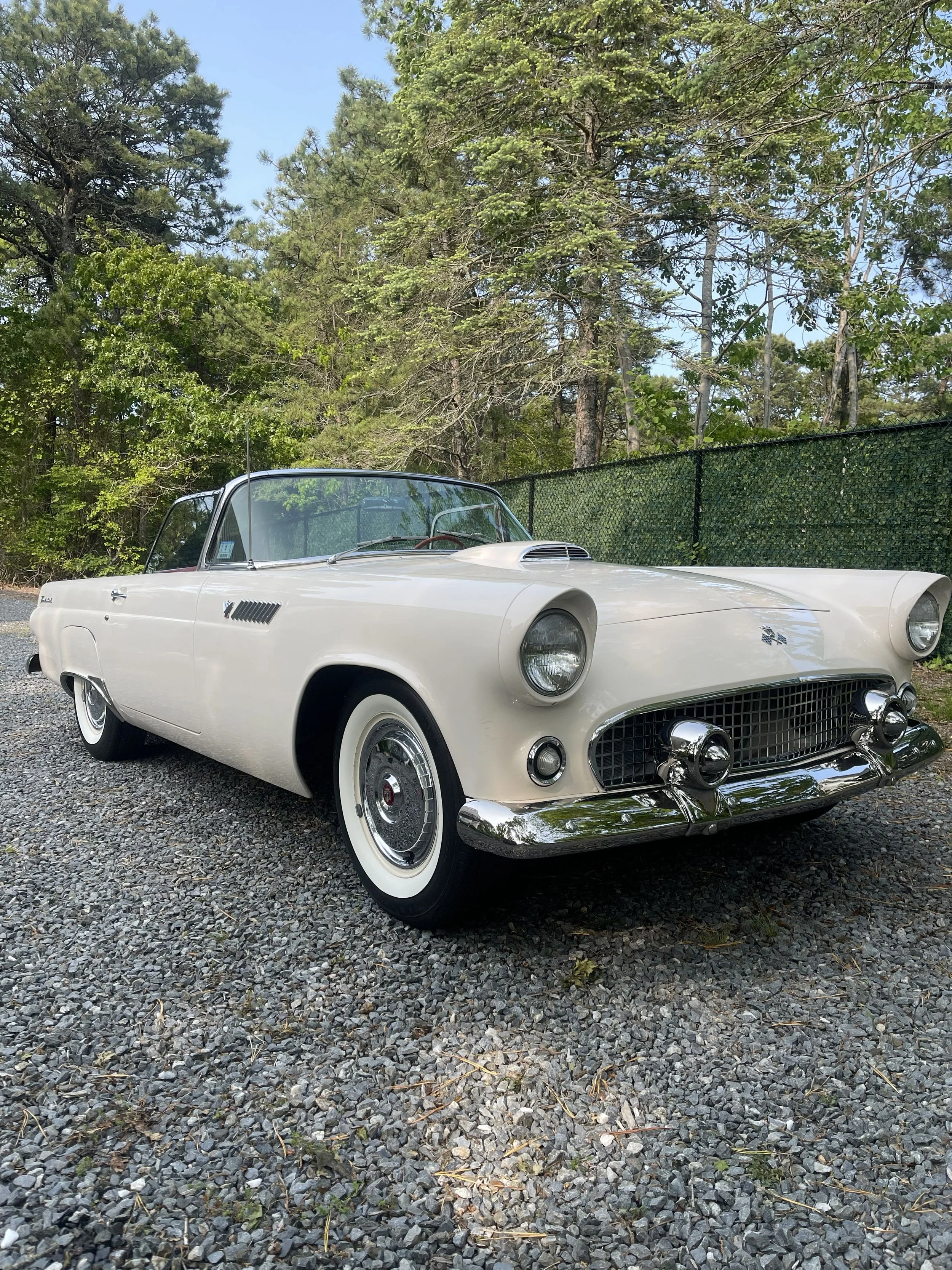 A vintage white convertible car parked on a gravel surface with a backdrop of trees and a black fence.