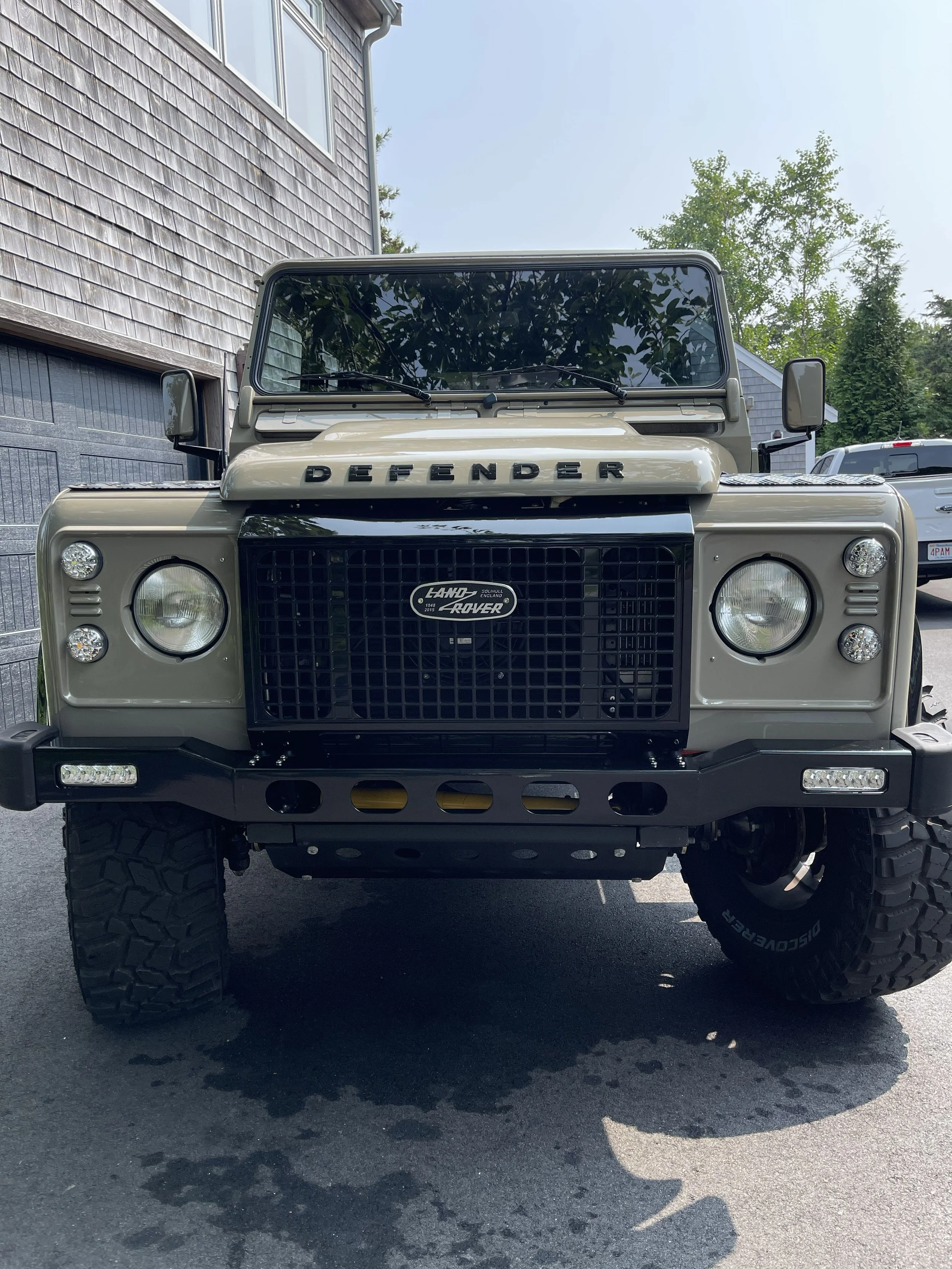 Front view of a beige Land Rover Defender with large tires, parked in front of a building with shingle siding, trees and another vehicle in the background.