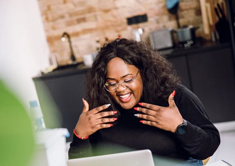 A woman with curly hair, glasses, and a watch, smiling and holding her hands near her face while looking at a laptop in a modern kitchen.