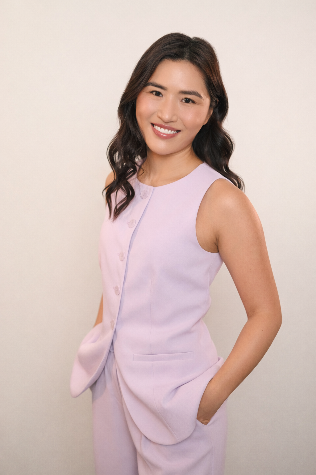 Young woman smiling, posing in a sleeveless light purple outfit against a plain background.