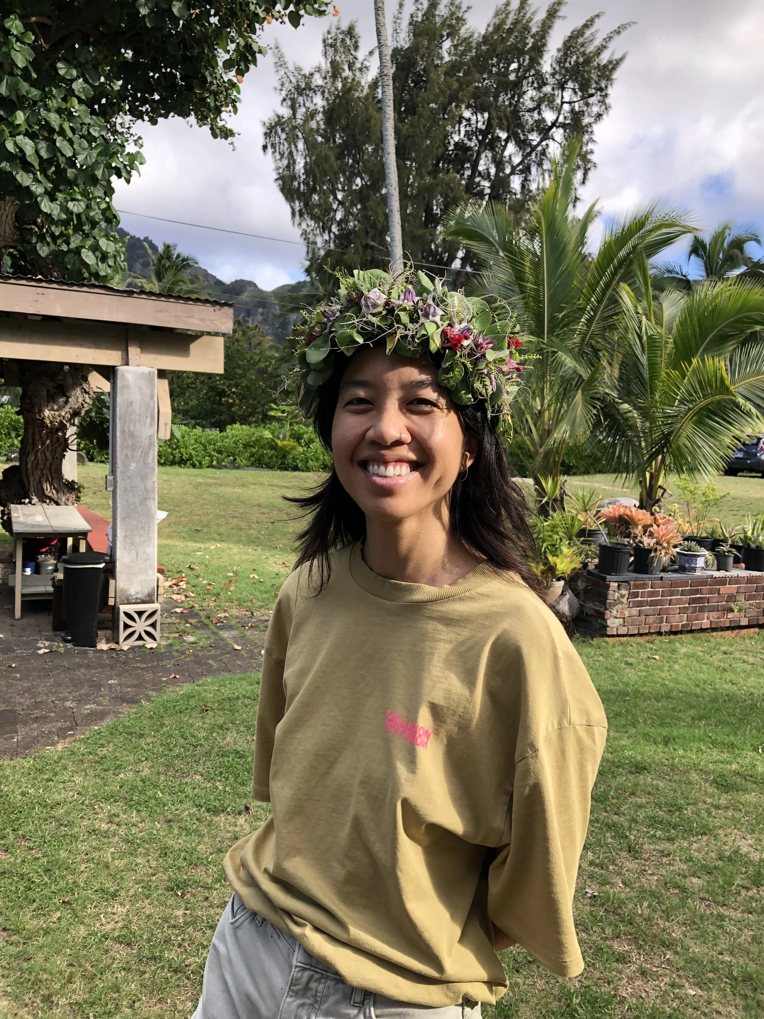 A woman with black hair wearing a flower crown outside standing on grass.