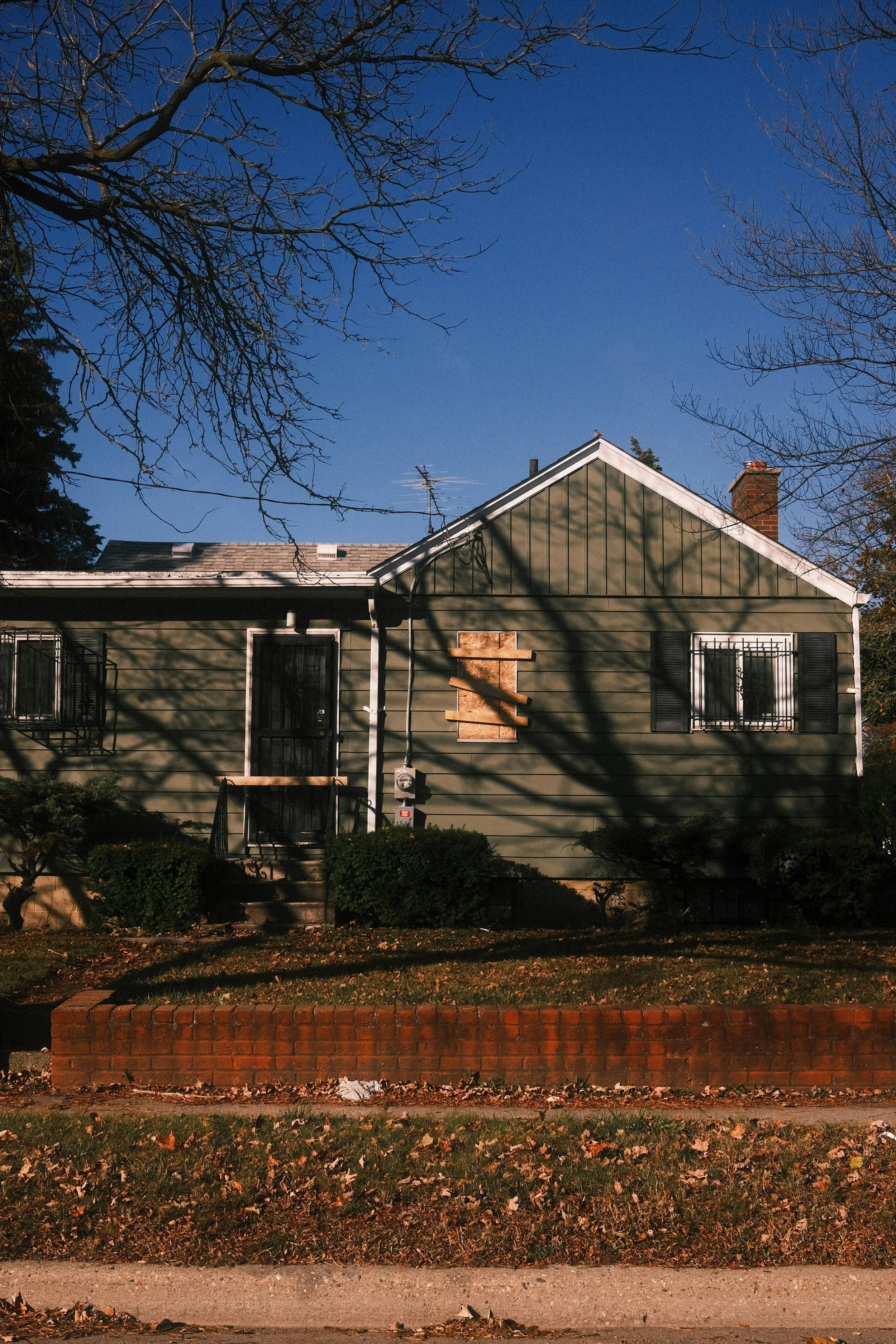 A house with dark green siding and black shutters, boarded up windows, and a boarded-up door, with leafless trees casting shadows on it and a clear blue sky in the background.