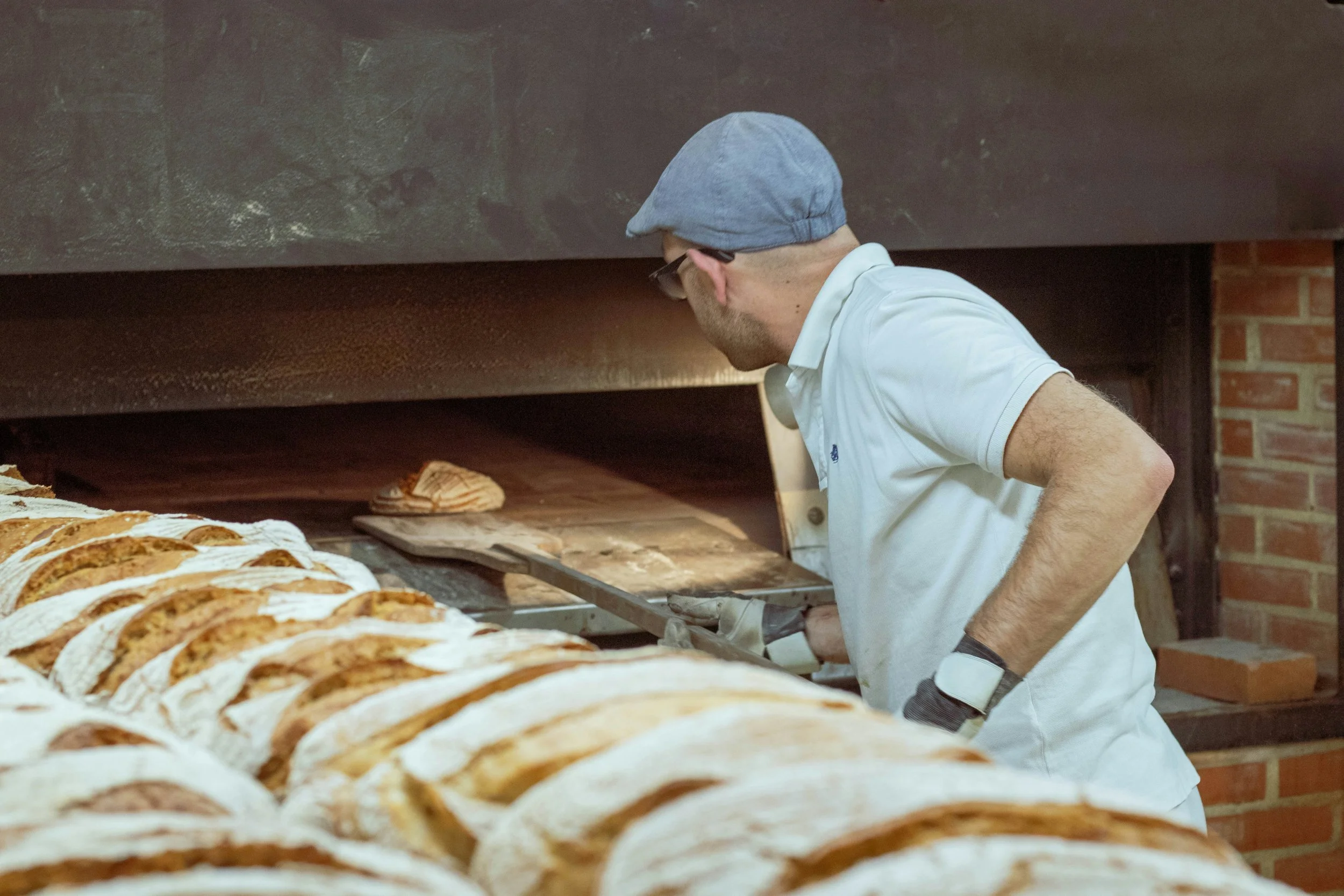 A man in a white shirt and gray cap baking bread in a wood-fired oven.