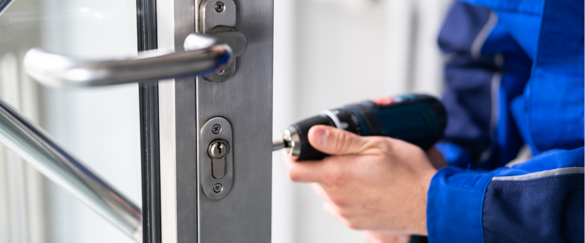 Person in blue work uniform using a cordless drill to install or fix a lock on a glass door.