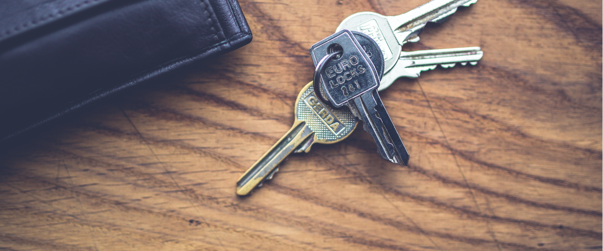 A set of keys on a wooden surface near a black wallet.