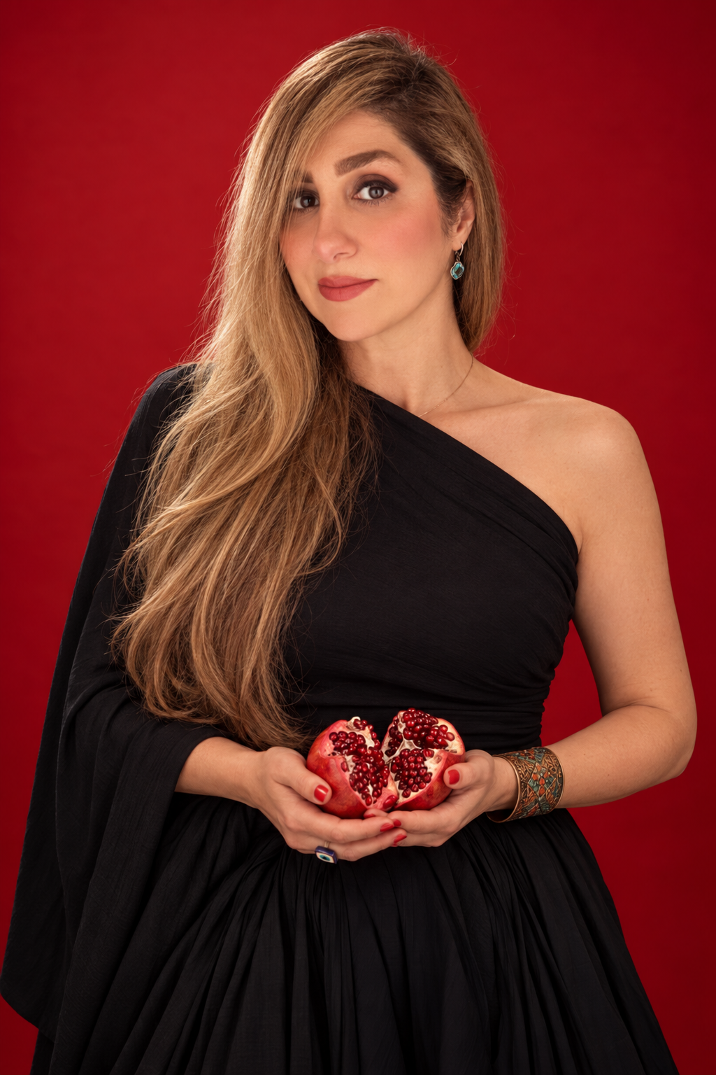 02 · Pomegranate Presence
An asymmetric black outfit and statement cuff against saturated red seamless. Holding an opened pomegranate at the waist, with steady eye contact and relaxed shoulders, communicates cultural connection and contemporary confi