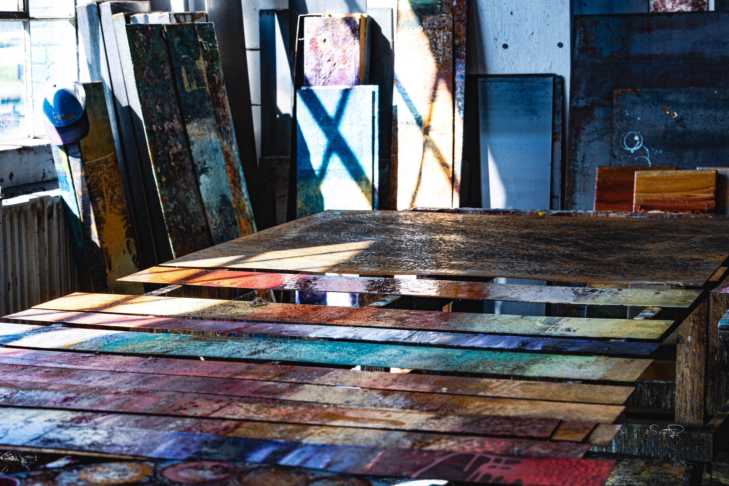 Window light casts diagonal shadows across colorful abstract steel paintings arranged on worktables in Mike Elsass’s Dayton studio.