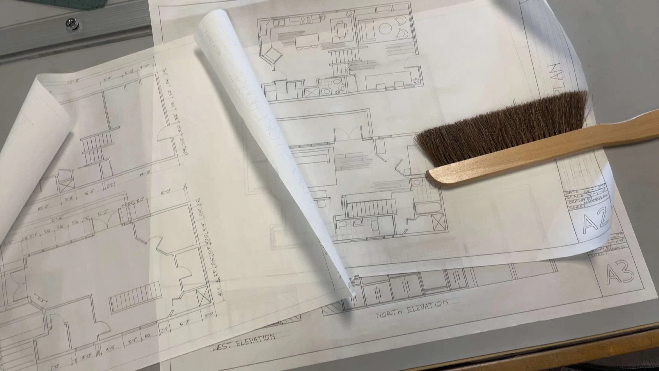 Construction or architectural blueprints spread out on a table, with a paintbrush and a floor plan diagram on top.