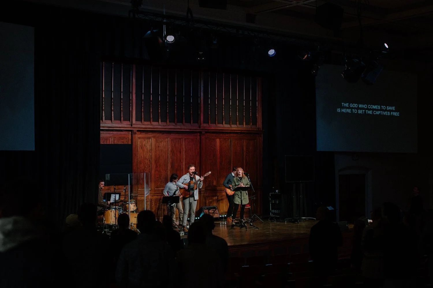 A band performs on stage in a church or auditorium, with a drummer, two guitarists, and a female vocalist, while people in the audience worship and listen; lyrics are projected on a screen to the right.