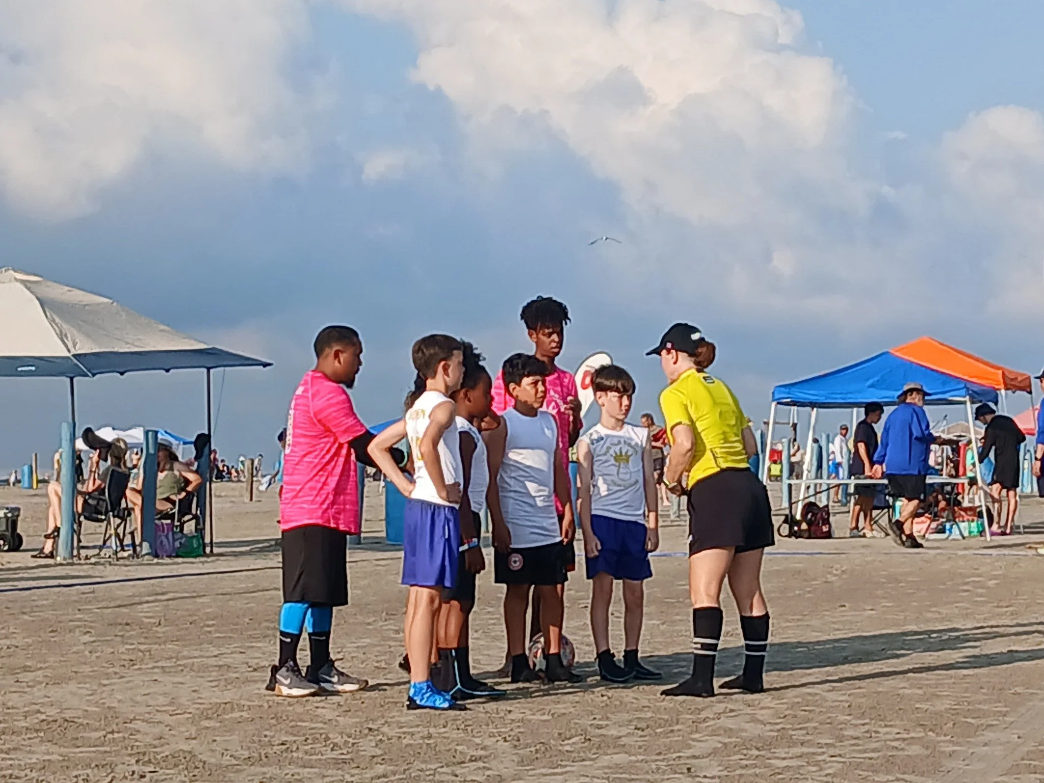 A group of young boys wearing sports uniforms are standing in a line on a sandy beach, listening to a referee in a yellow shirt and black shorts, who appears to be giving instructions or a speech. There are tents, umbrellas, and people in the background enjoying the beach.