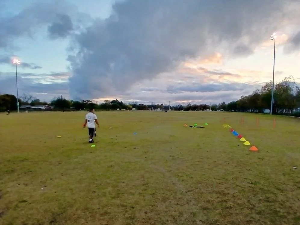A football practice field during sunset with cones and training equipment, a person walking, and floodlights illuminating the field.