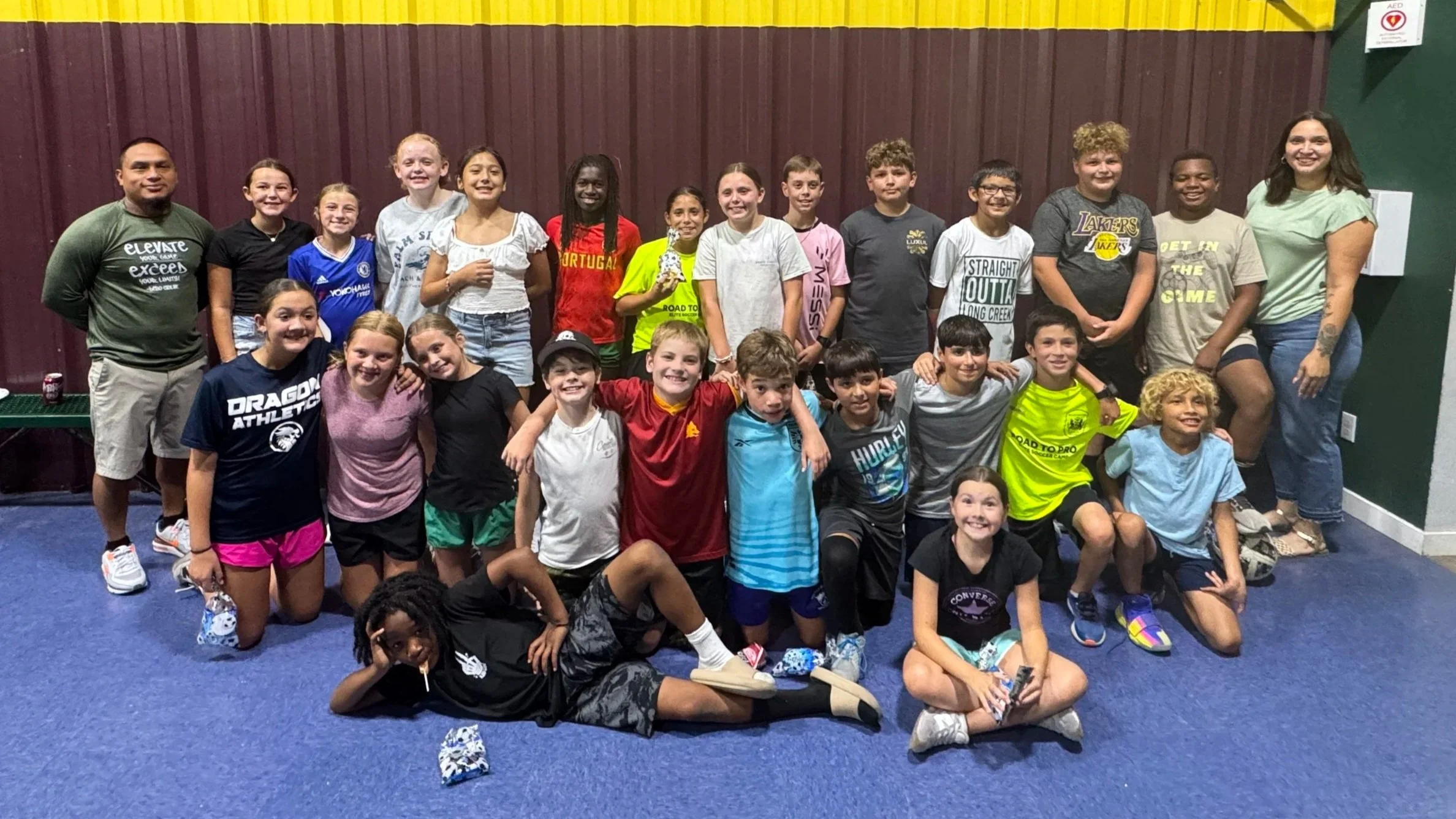 Group of children and two adults posing together indoors in front of a maroon wall, some children kneeling or sitting on the floor, others standing behind, all smiling.