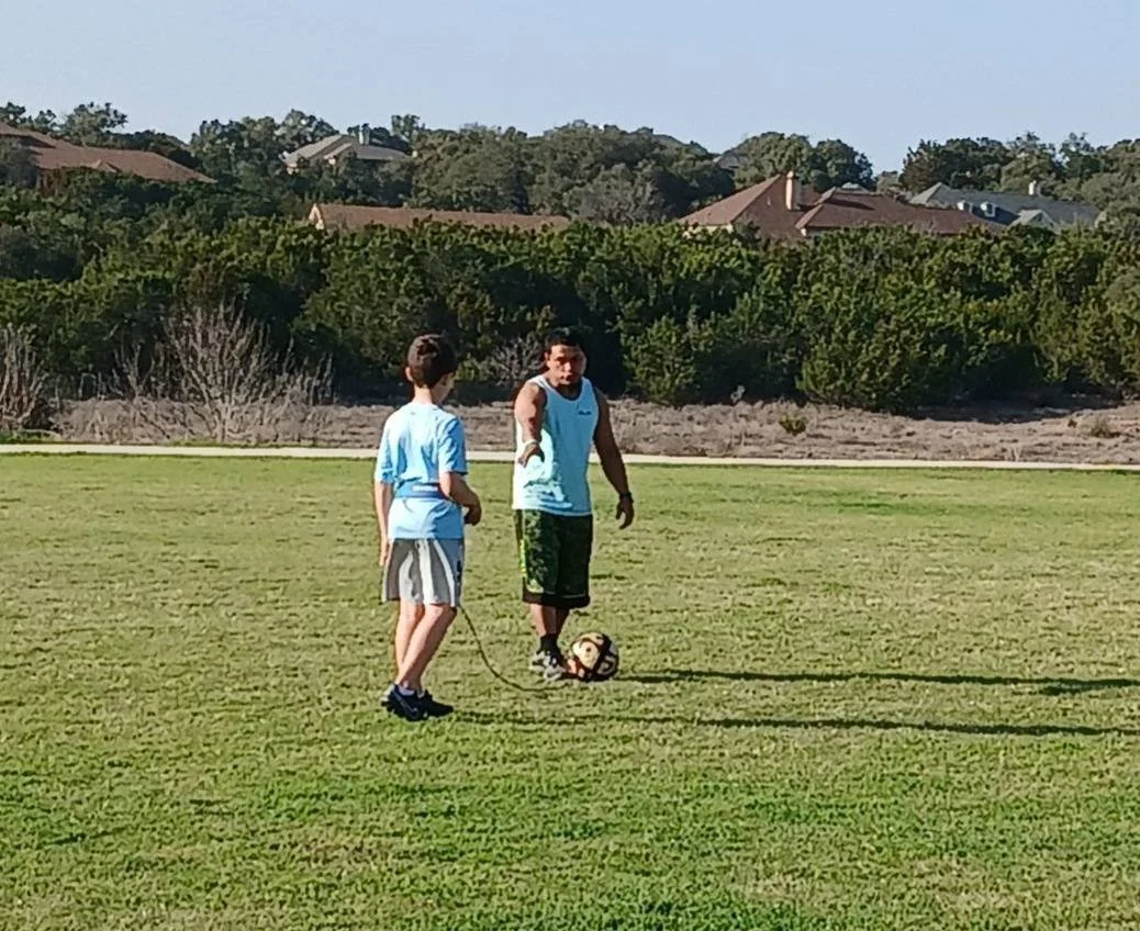 Two boys on a grassy field, one holding a jump rope and the other standing with a soccer ball, with houses and trees in the background.